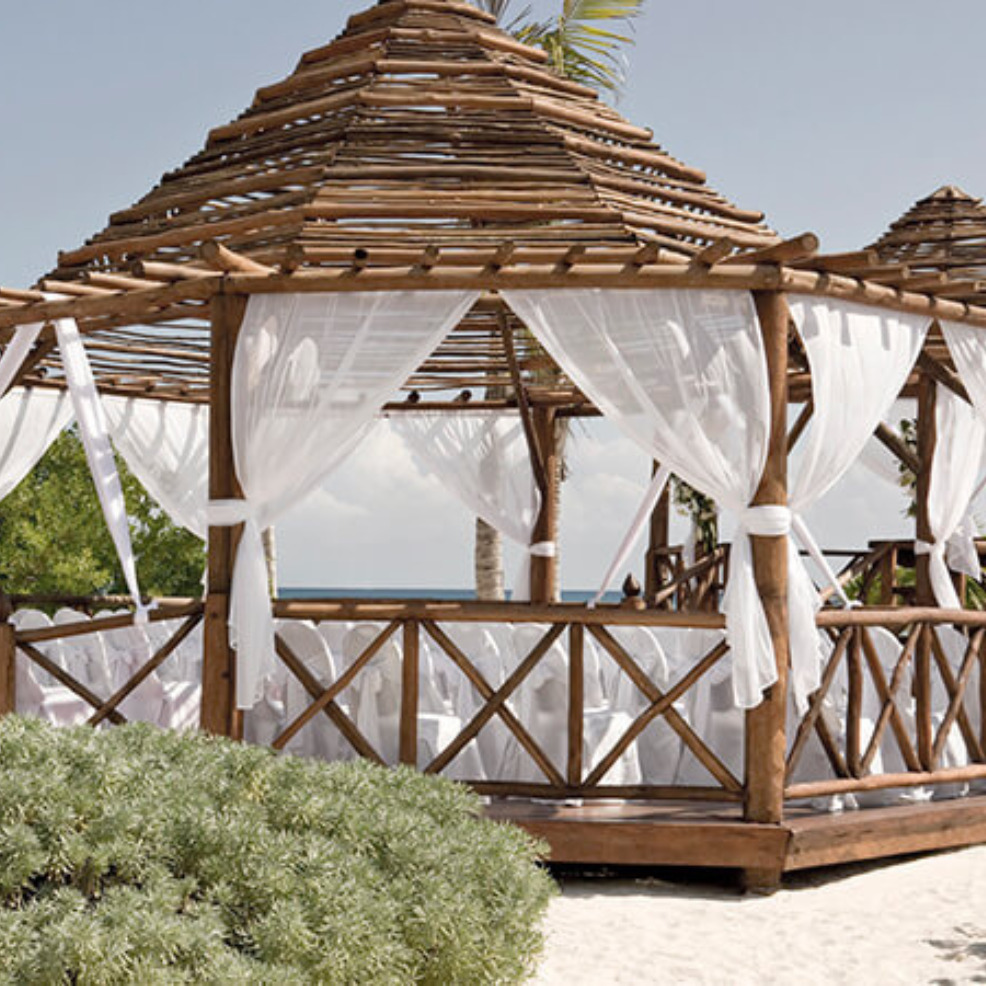Wooden beach cabana with white sheer curtains on a sandy shore, perfect for a destination wedding, under a clear blue sky.