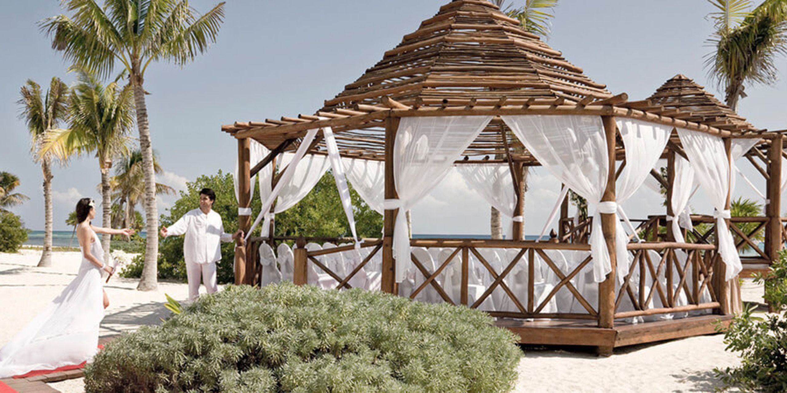 A couple in wedding attire stands apart on a sandy beach with a wooden gazebo decorated with white drapes in the background at their destination wedding.