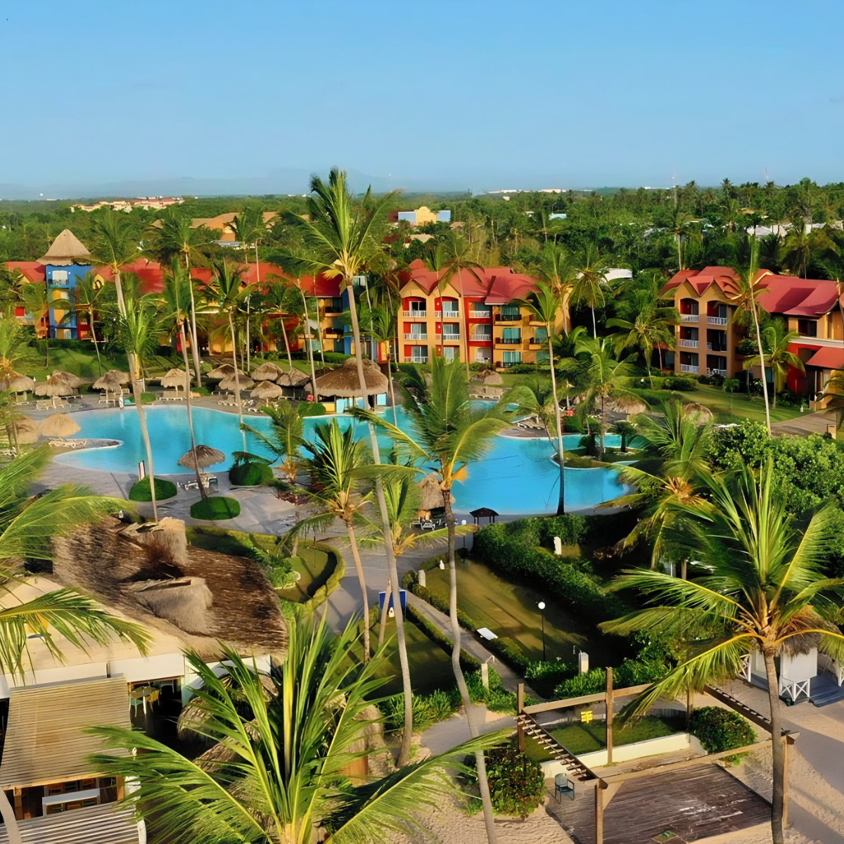 Aerial view of the Punta Cana Princess, a tropical resort popular for destination weddings, with vividly colored buildings, large swimming pools, and lush palm trees, located near the coast.
