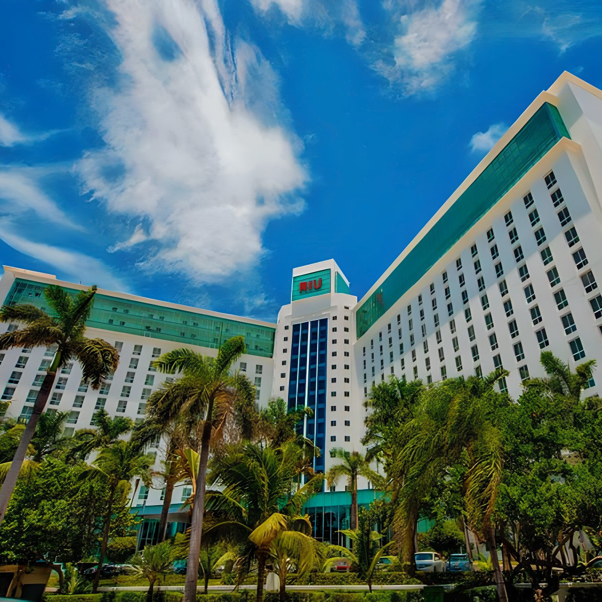 A vibrant image of the Hotel Riu Cancun building, a popular destination for weddings, under a bright blue sky with lush green palm trees in the foreground.