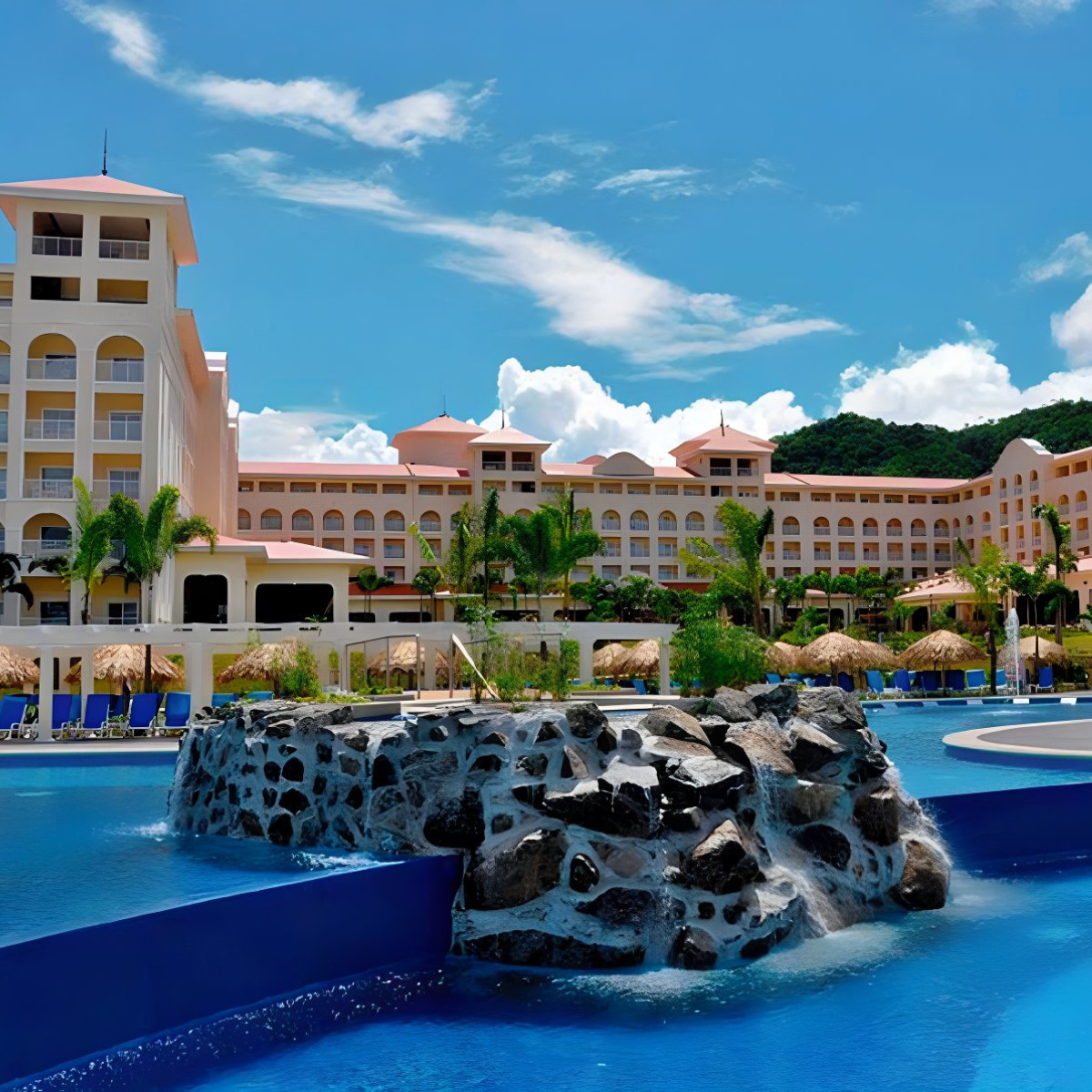 Hotel Riu Guanacaste, ideal for destination weddings, with a large swimming pool and decorative rock feature in the foreground, surrounded by a multi-story pink building under a clear sky.