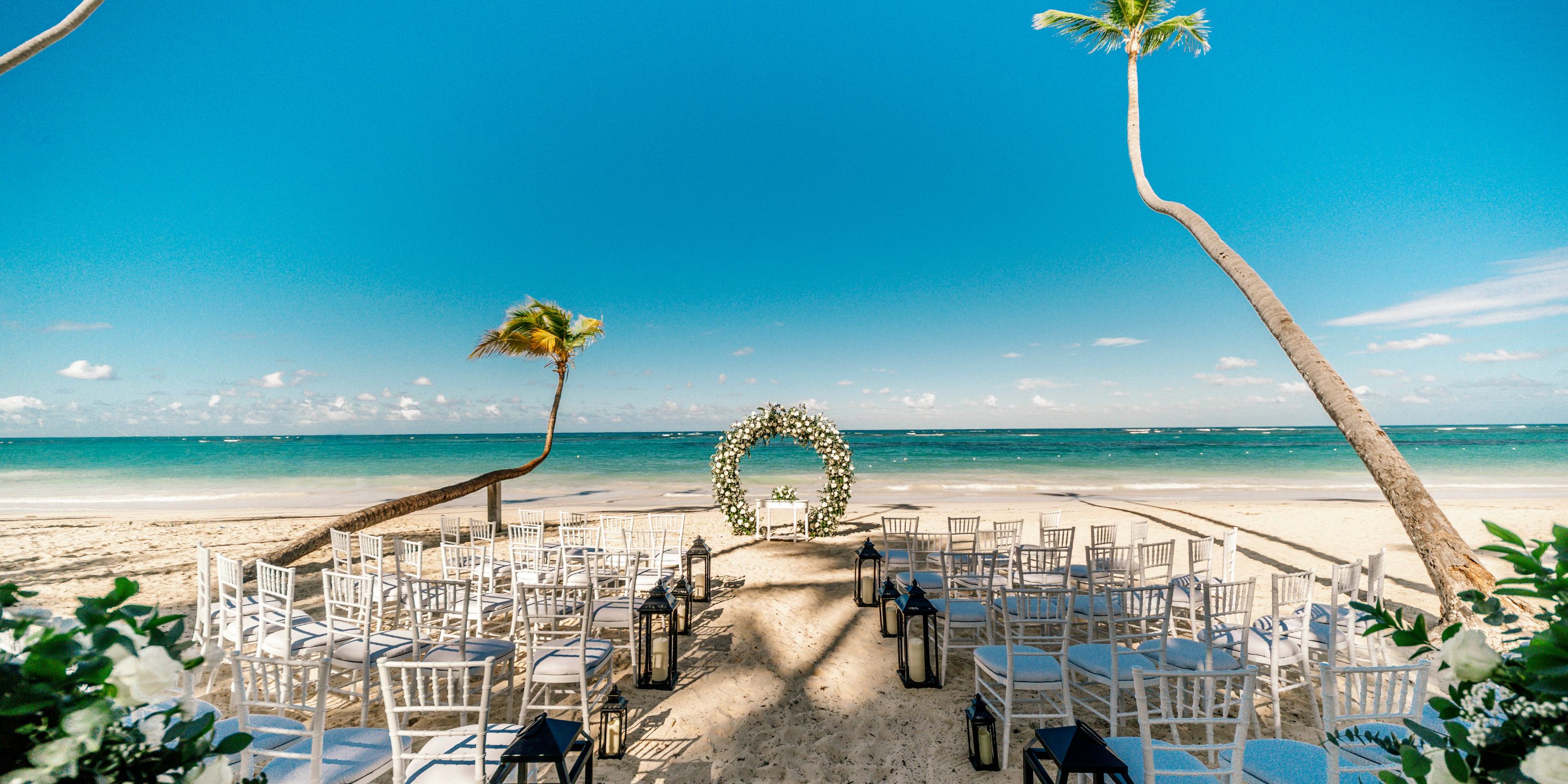 White chairs set for a destination wedding face a floral arch on the sandy beach at Hotel Riu Bambu.