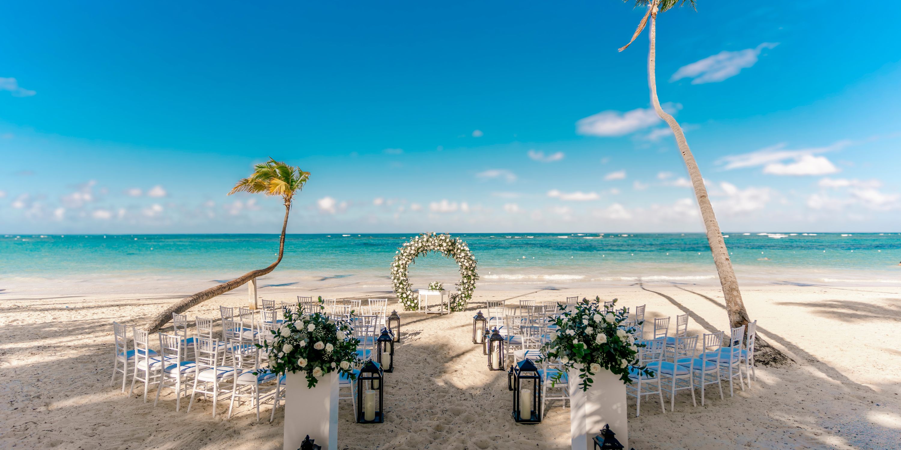 Beachfront chairs and a floral arch set for a destination wedding at Hotel Riu Palace Bavaro under blue sky.