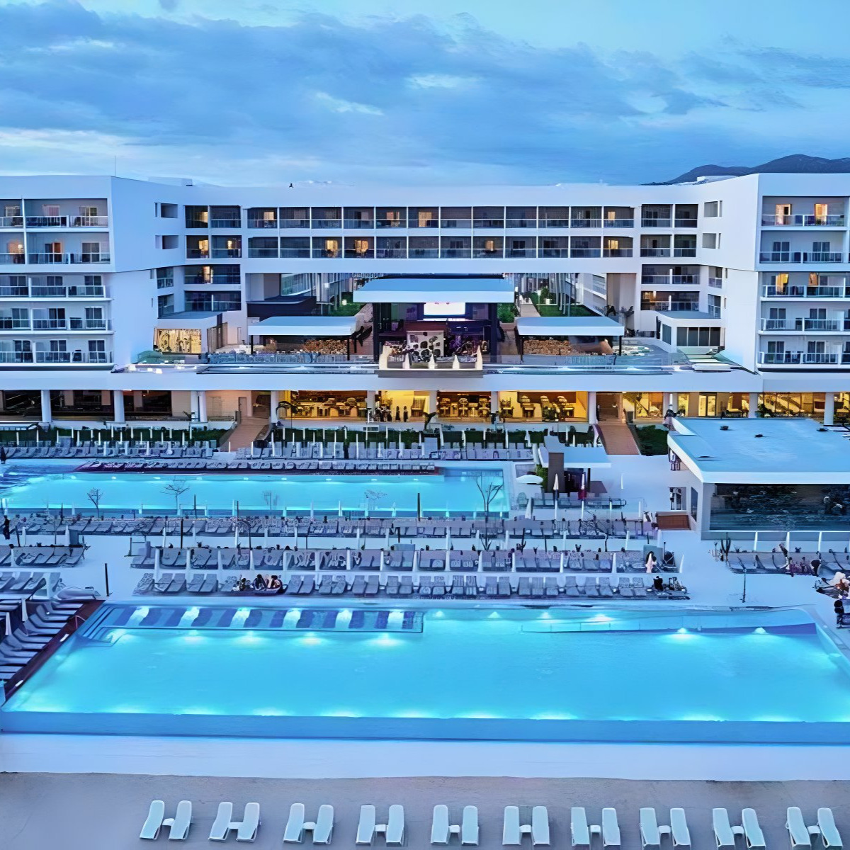 A luxury destination wedding Hotel Riu Palace Baja California with bright blue lit pools in the evening, surrounded by white lounge chairs and a modern white building structure.