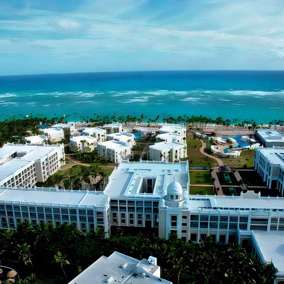 Aerial view of Hotel Riu Palace Bavaro, popular for destination weddings, with white buildings and lush greenery, overlooking a turquoise ocean.