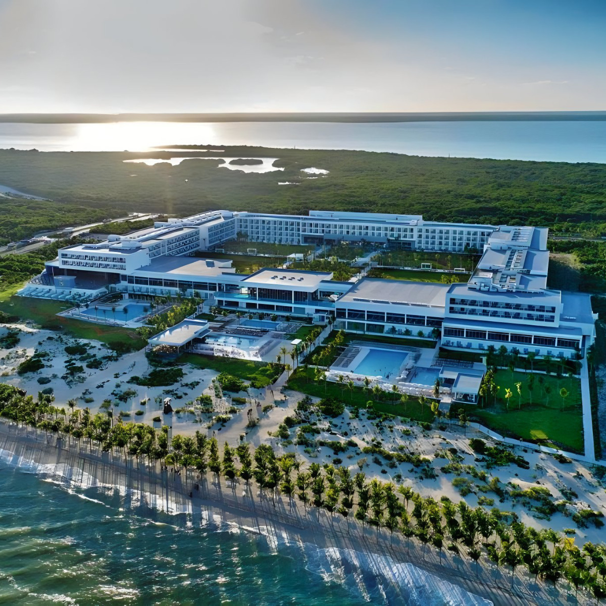 Aerial view of Hotel Riu Palace Costa Mujeres, a large, modern resort complex beside a beach, ideal for destination weddings, with surrounding vegetation at sunset.