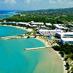 Aerial view of Hotel Riu Palace Jamaica, a tropical destination weddings resort with white buildings along a curving sandy beach, surrounded by clear turquoise waters and lush greenery.
