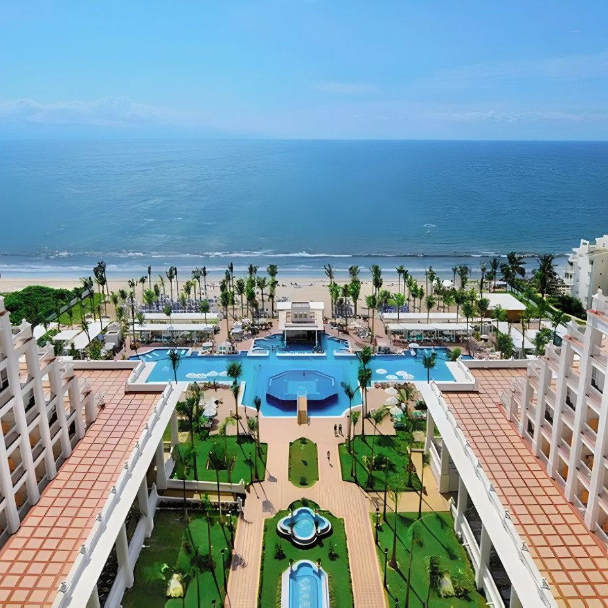 Aerial view of Hotel Riu Palace Pacifico, ideal for a destination wedding, with swimming pools and palm trees, situated between a white sandy beach and hotel buildings.