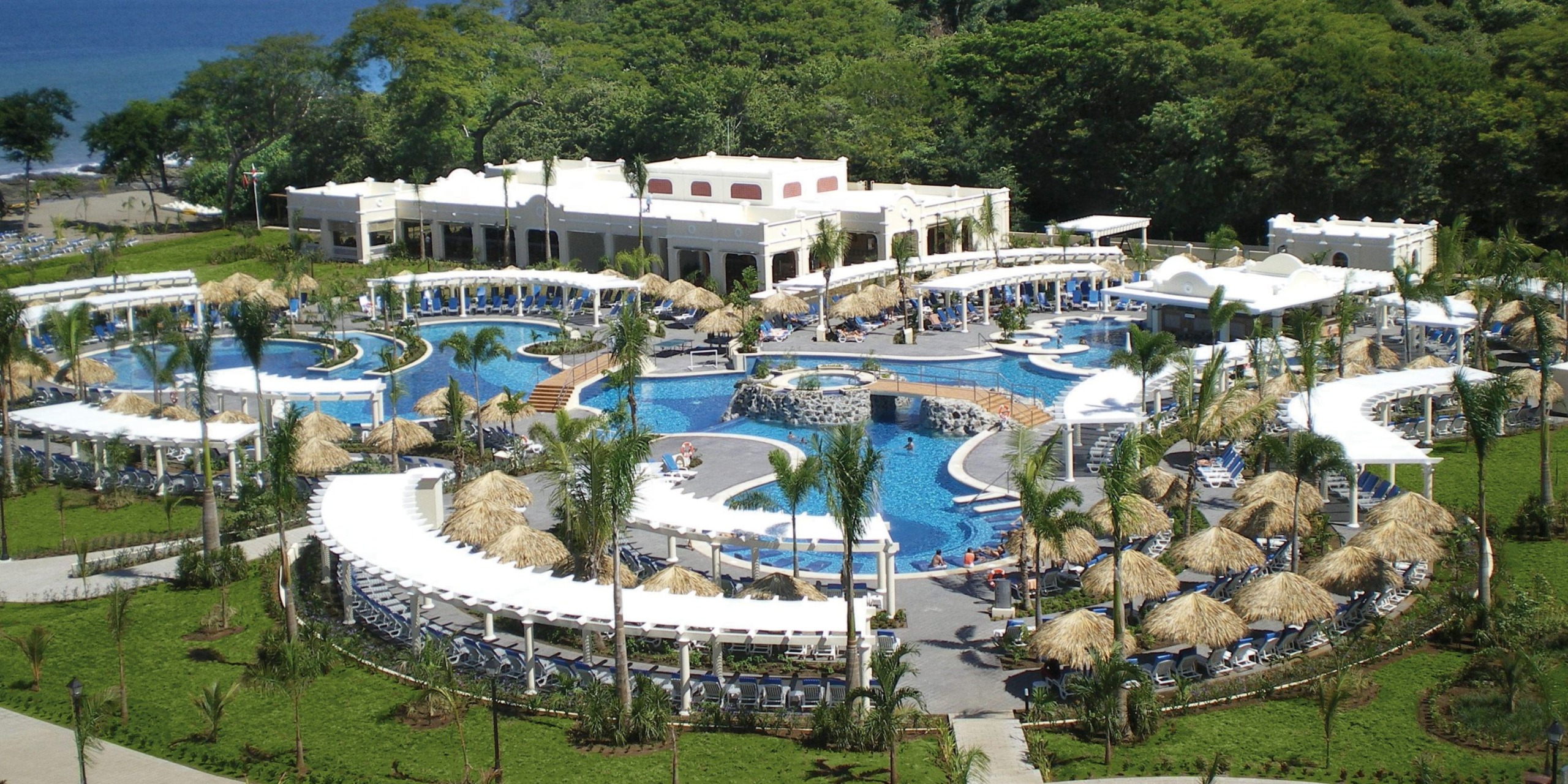 Aerial view of the Hotel Riu Guanacaste, featuring a large, winding pool encircled by palm trees, lounge chairs, and thatched umbrellas. A central building is nestled among lush greenery.