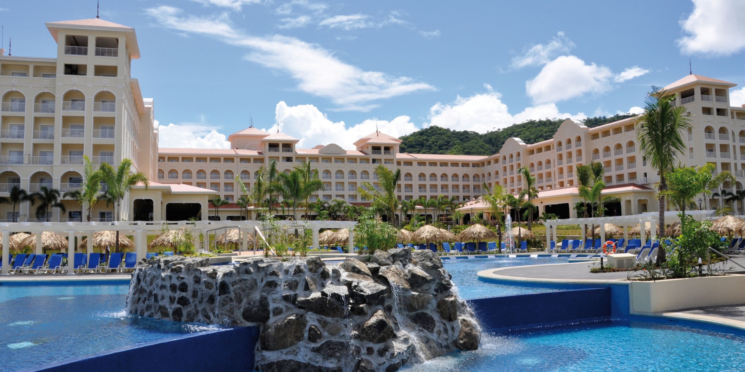 The Hotel Riu Guanacaste offers a luxurious experience with multiple balconies overlooking a spacious pool area, featuring a stunning stone waterfall, all surrounded by lush palm trees under a clear blue sky.