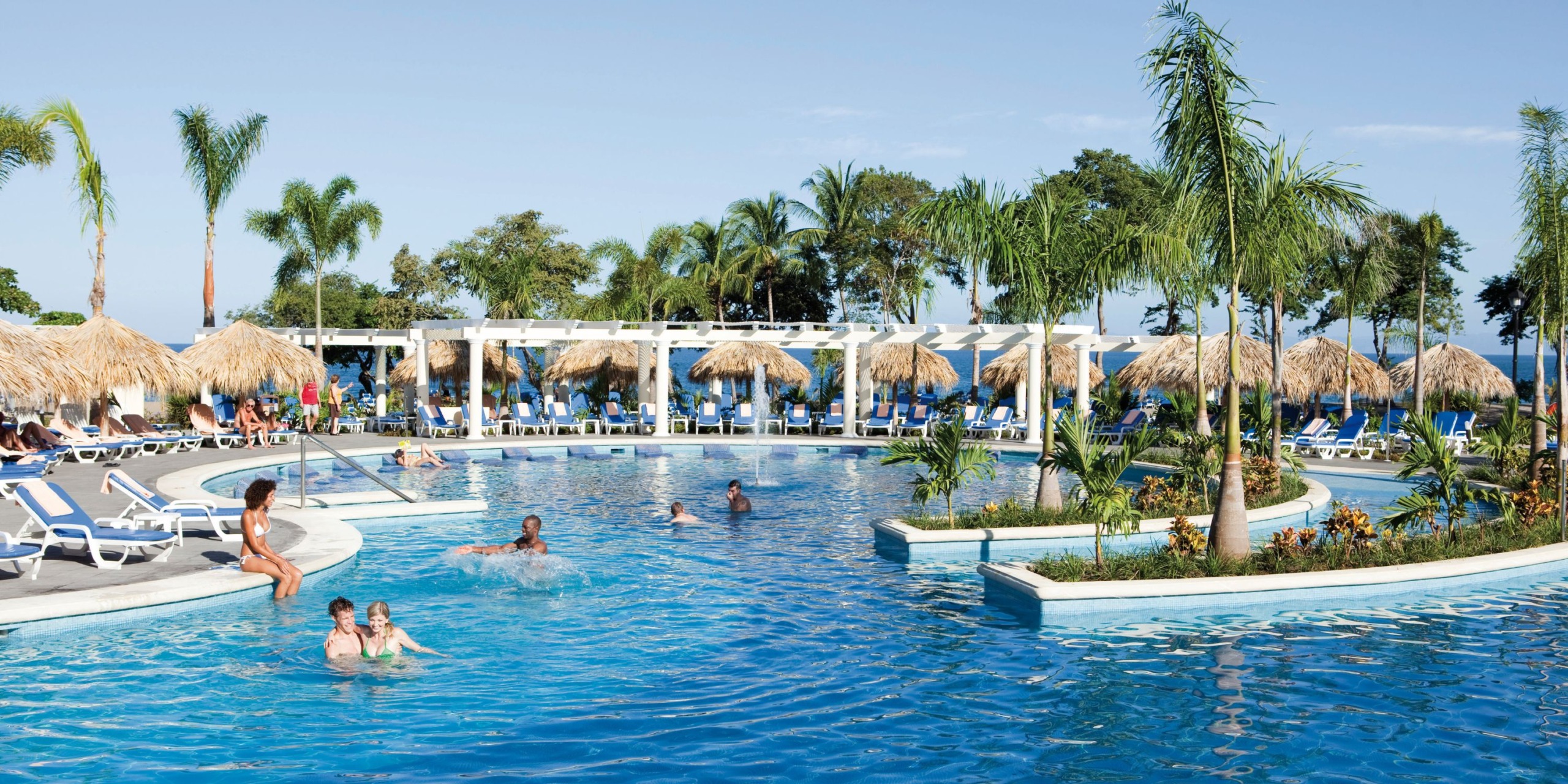 A large outdoor swimming pool at Hotel Riu Guanacaste features people swimming and sunbathing, surrounded by palm trees and straw umbrellas that create a tropical atmosphere.