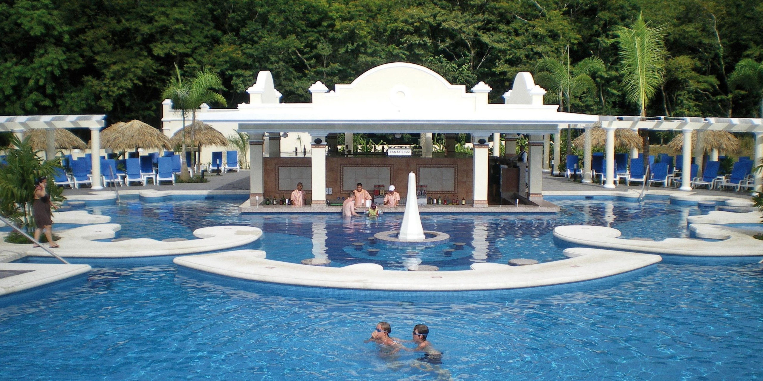 The Hotel Riu Guanacaste features an outdoor pool with a central white fountain. A poolside bar surrounded by lounge chairs and lush greenery invites relaxation. Two guests are enjoying the water while others unwind at the bar.