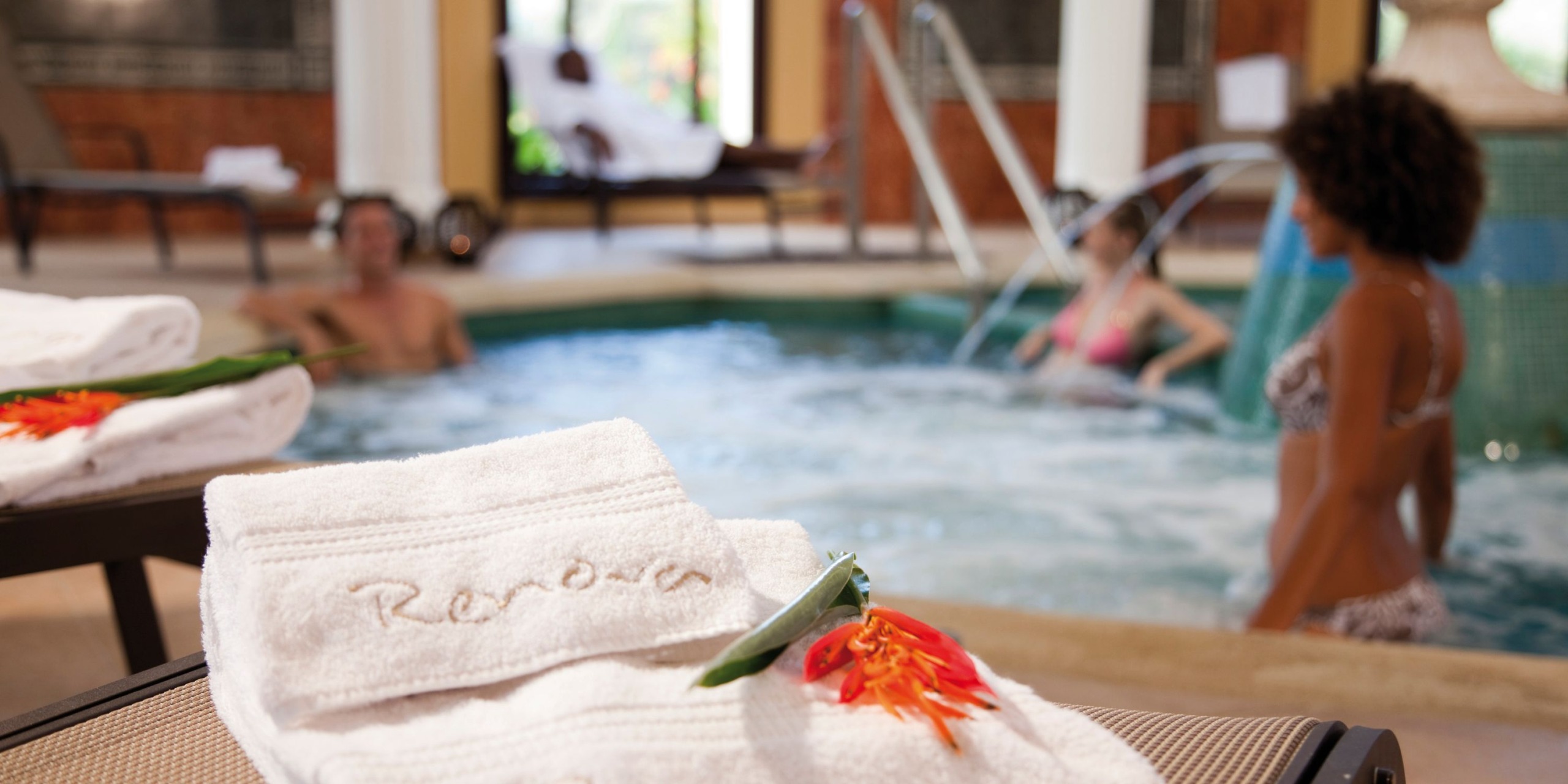 Towels featuring a floral design draped over a lounge chair in front of an indoor hot tub at Hotel Riu Guanacaste, where two people are enjoying their relaxation.