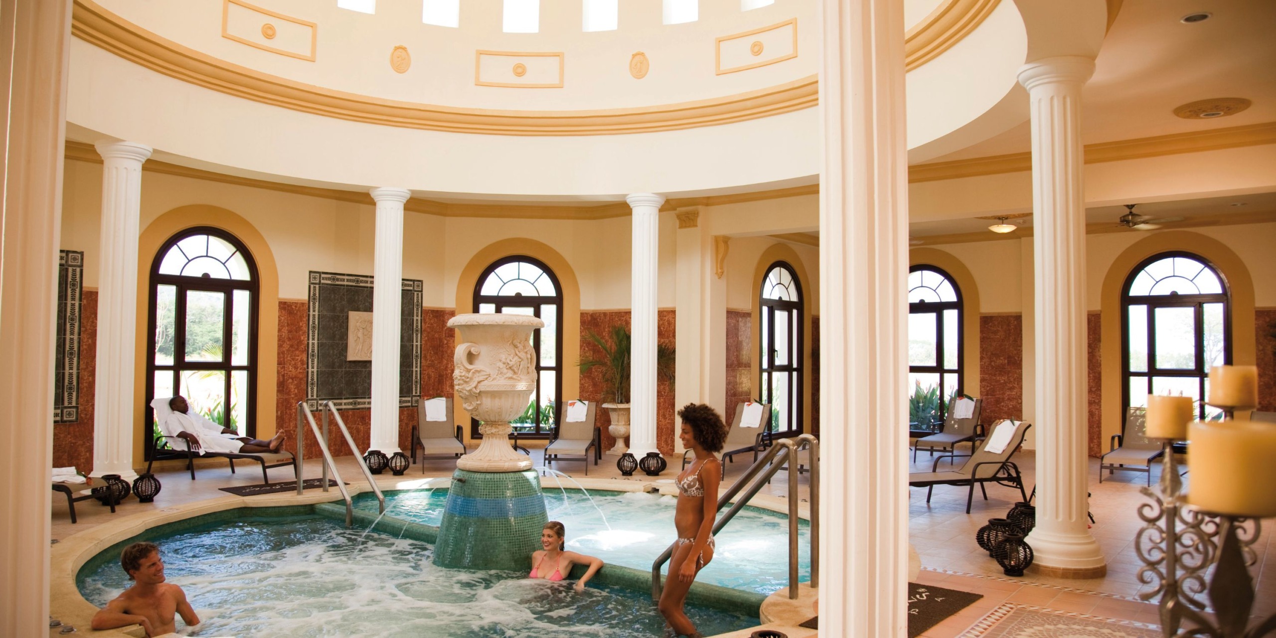 Guests unwinding in the opulent indoor spa pool at Hotel Riu Guanacaste, surrounded by soaring high ceilings, elegant columns, and plush lounge chairs.