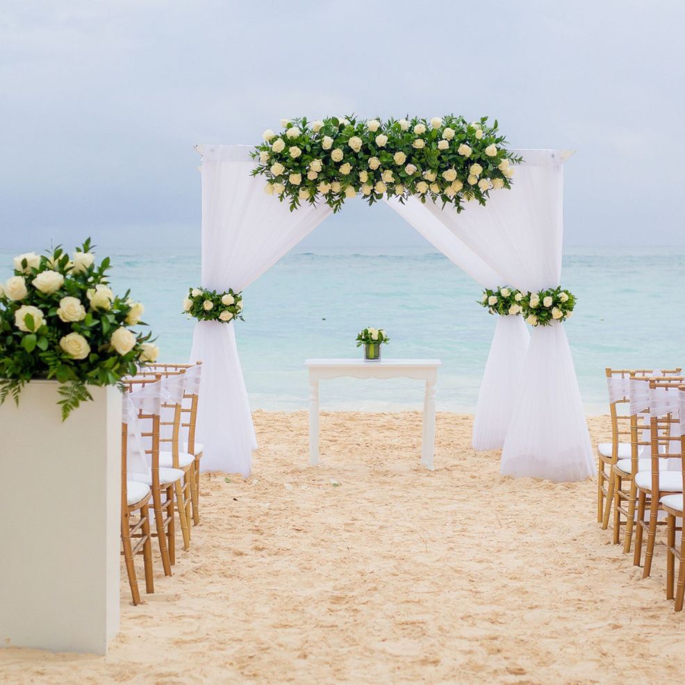 Destination wedding setup on the beach with white flowers, chairs, and an arch overlooking the ocean.
