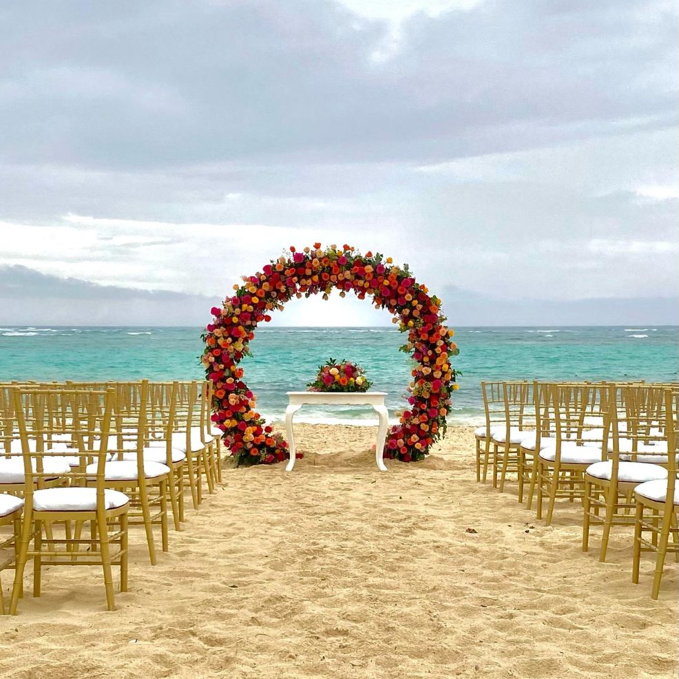 Destination wedding scene with a floral arch, white chairs, and table arranged by the seaside.