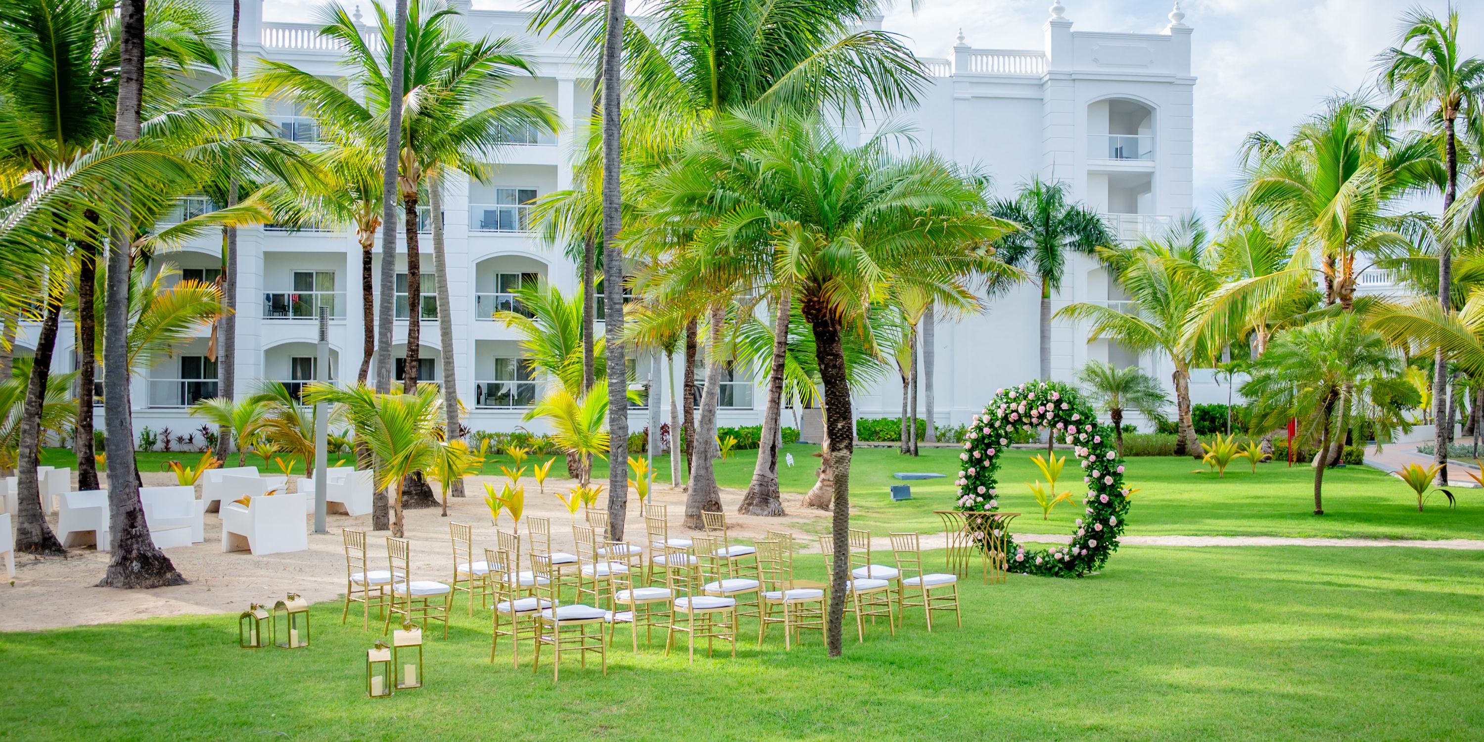 Destination wedding ceremony on a grassy lawn with gold chairs and a floral arch by a white building.