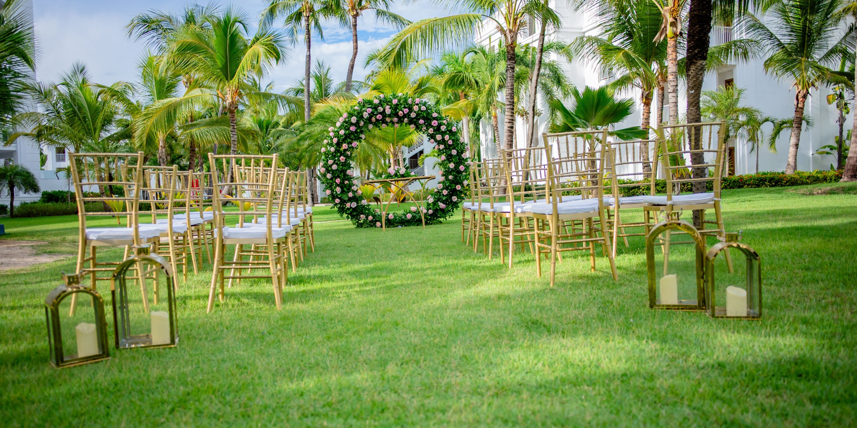 Destination wedding ceremony outdoors featuring gold chairs, a floral arch, and lanterns on lush grass.