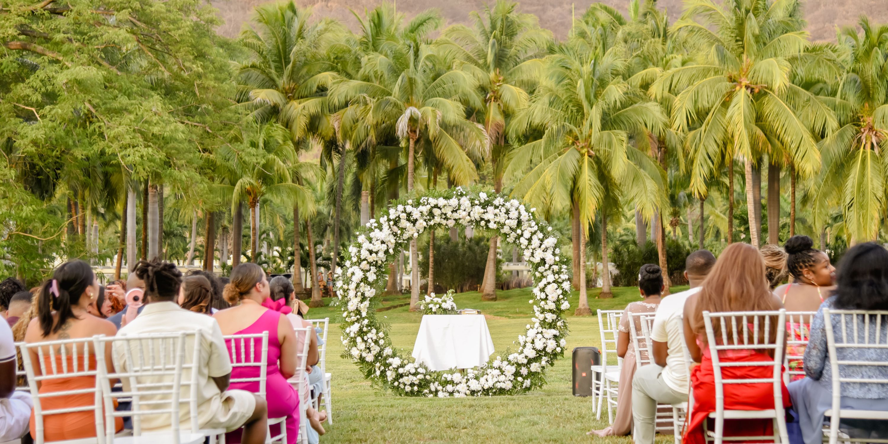 Destination wedding ceremony outdoors with guests in white chairs and a circular floral arch ahead.