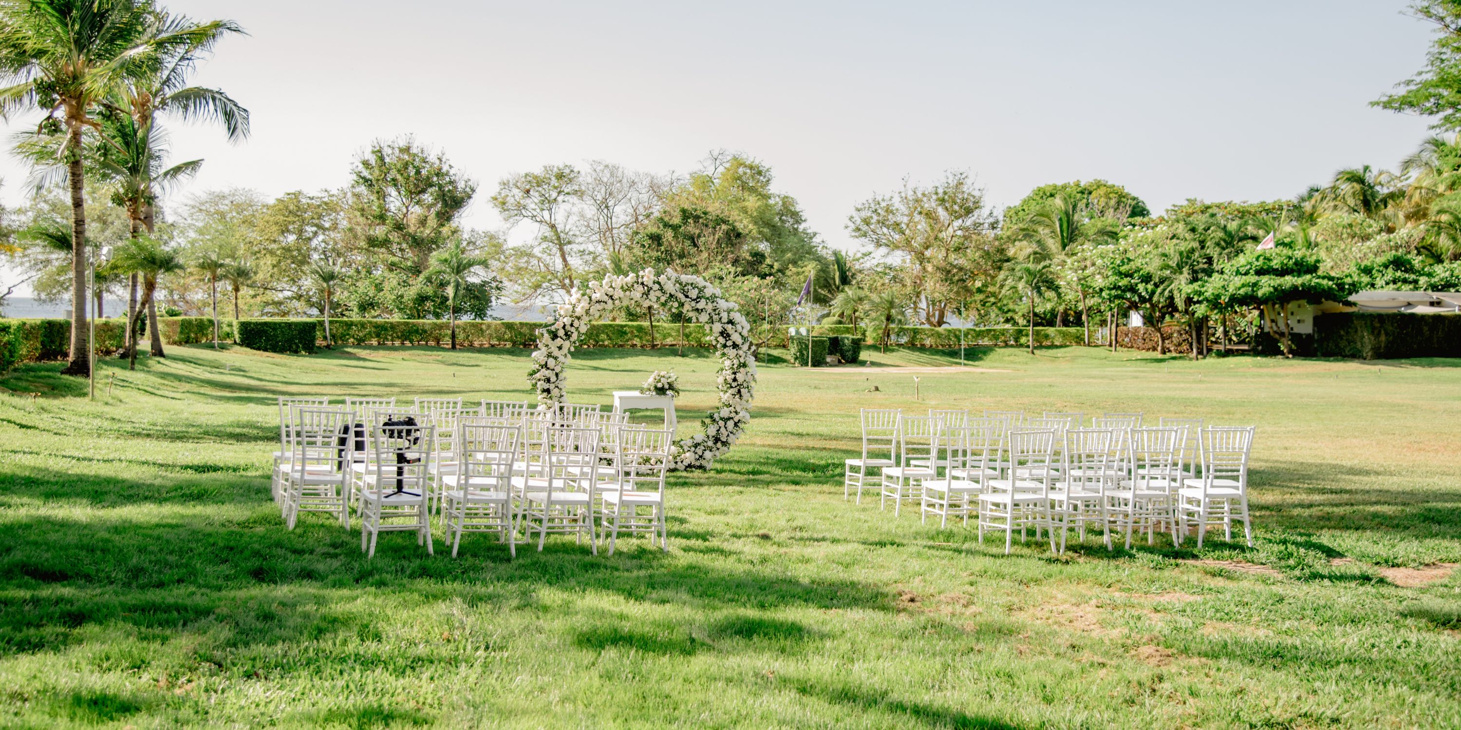Destination wedding ceremony featuring white chairs arranged on green grass with a circular floral arch.