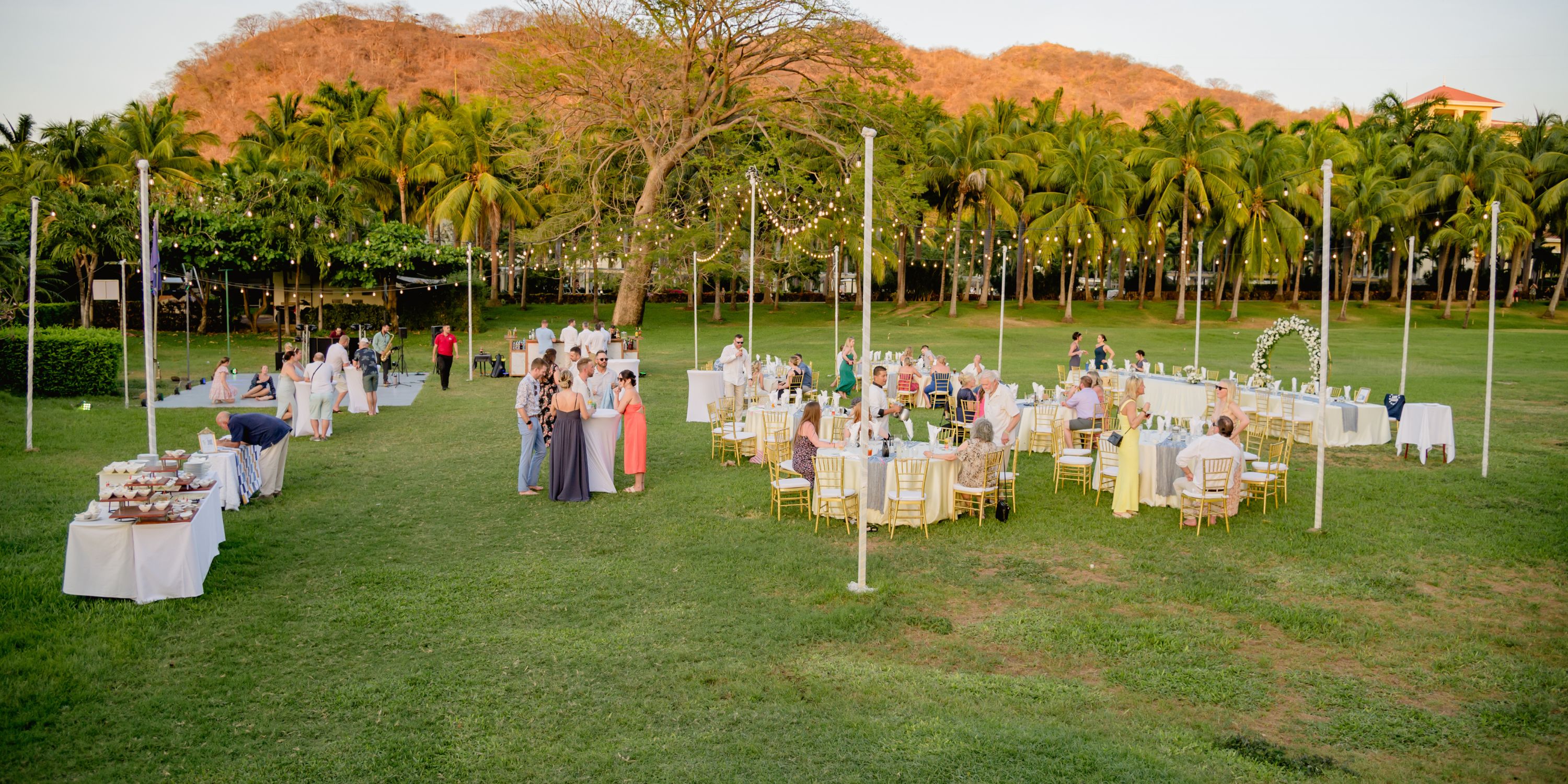 Destination wedding reception outdoors with guests at round tables on a lawn beneath string lights.