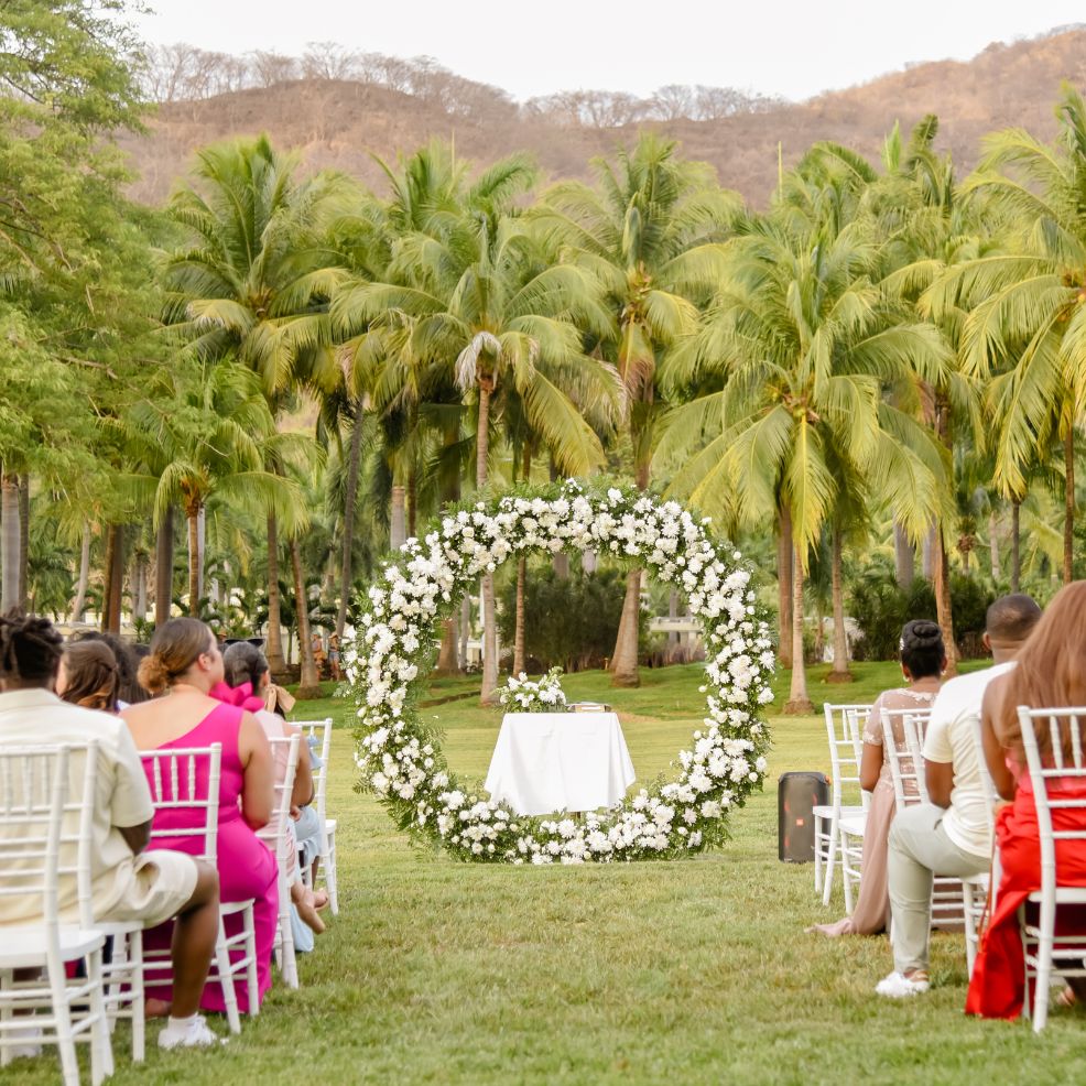 Destination wedding ceremony arrangement featuring white chairs and a round floral arch on green grass.