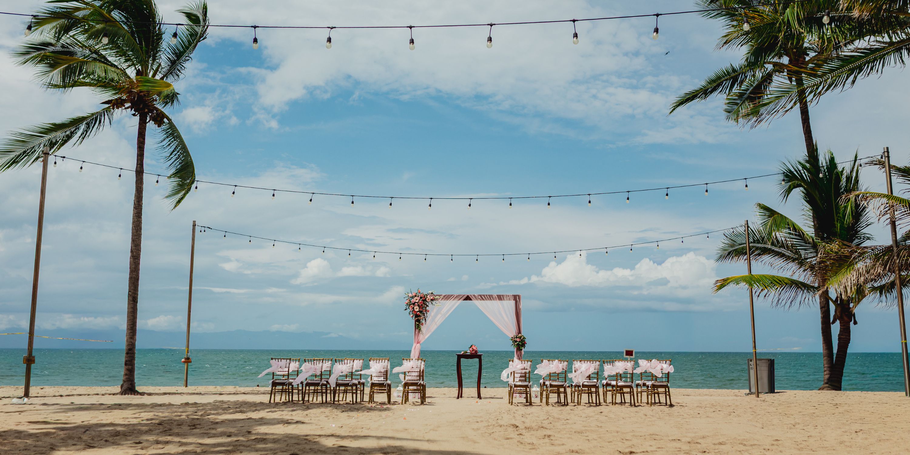 A destination wedding setup on the beach, with chairs and an arch facing the ocean beneath string lights.