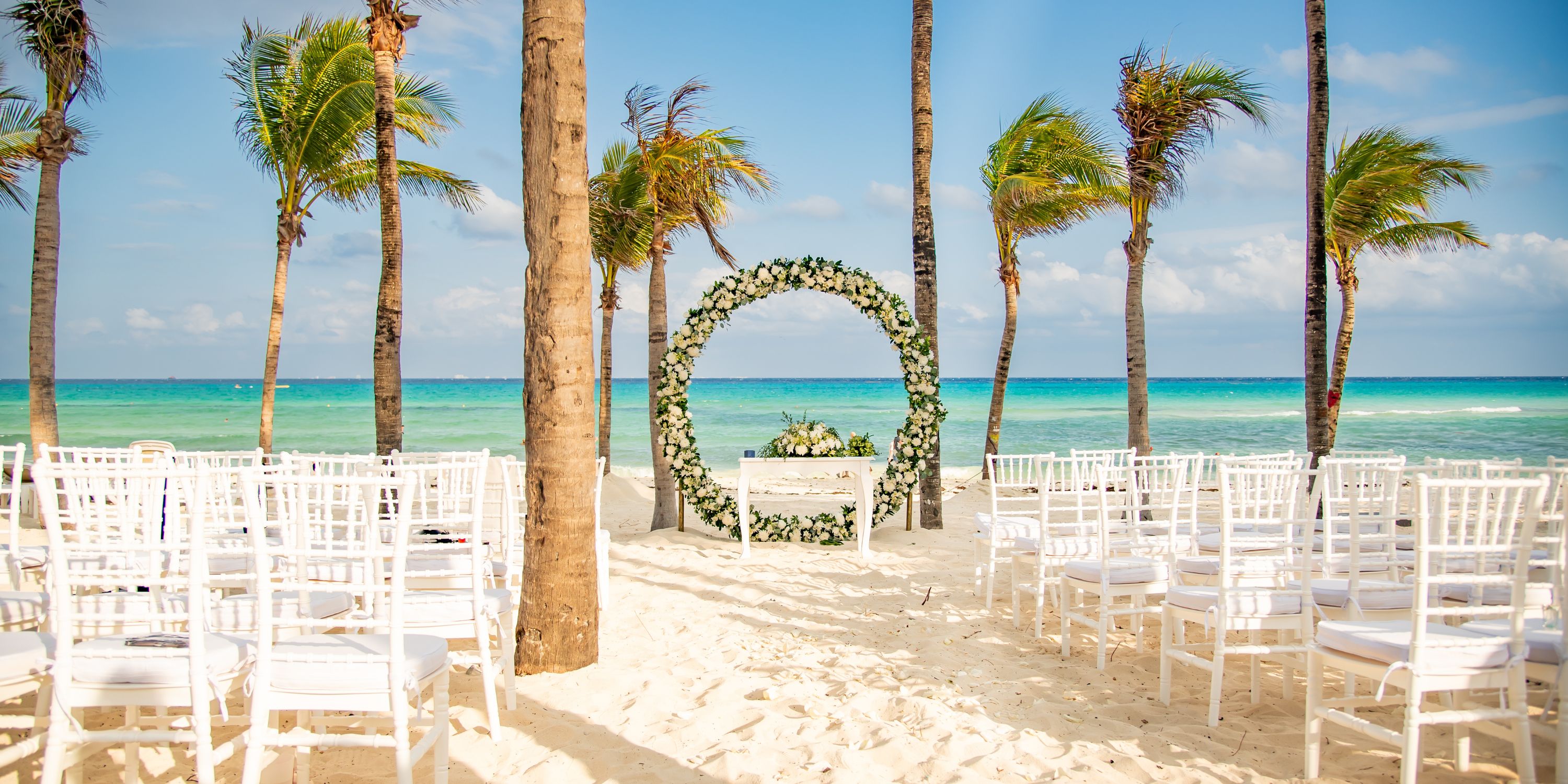 White chairs arranged for a destination wedding face a floral arch on the sandy beach at Hotel Riu Palace Riviera Maya.