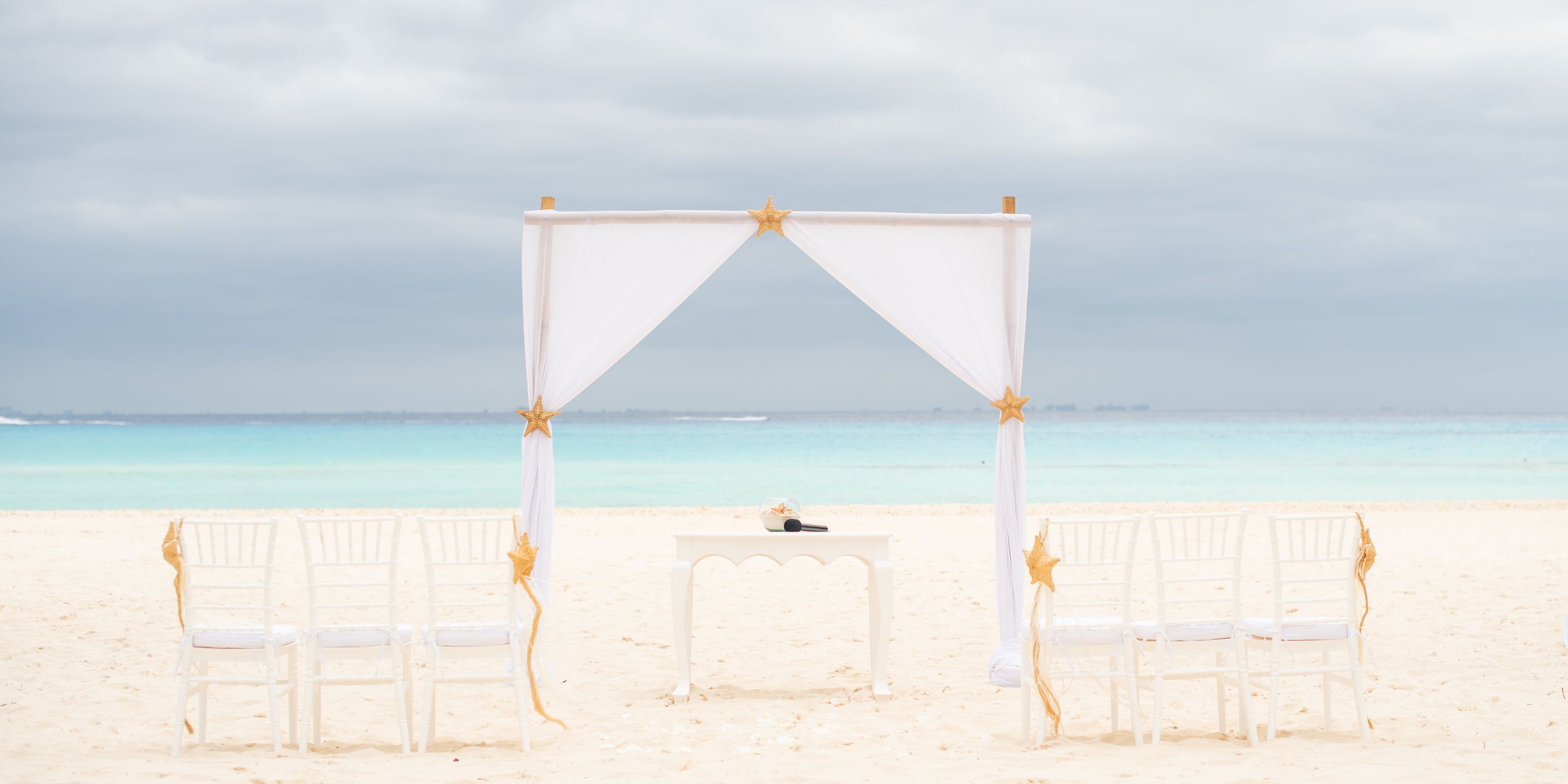 Destination wedding setup at Hotel Riu Yucatan: arch and chairs on sandy beach, sea and cloudy sky behind.