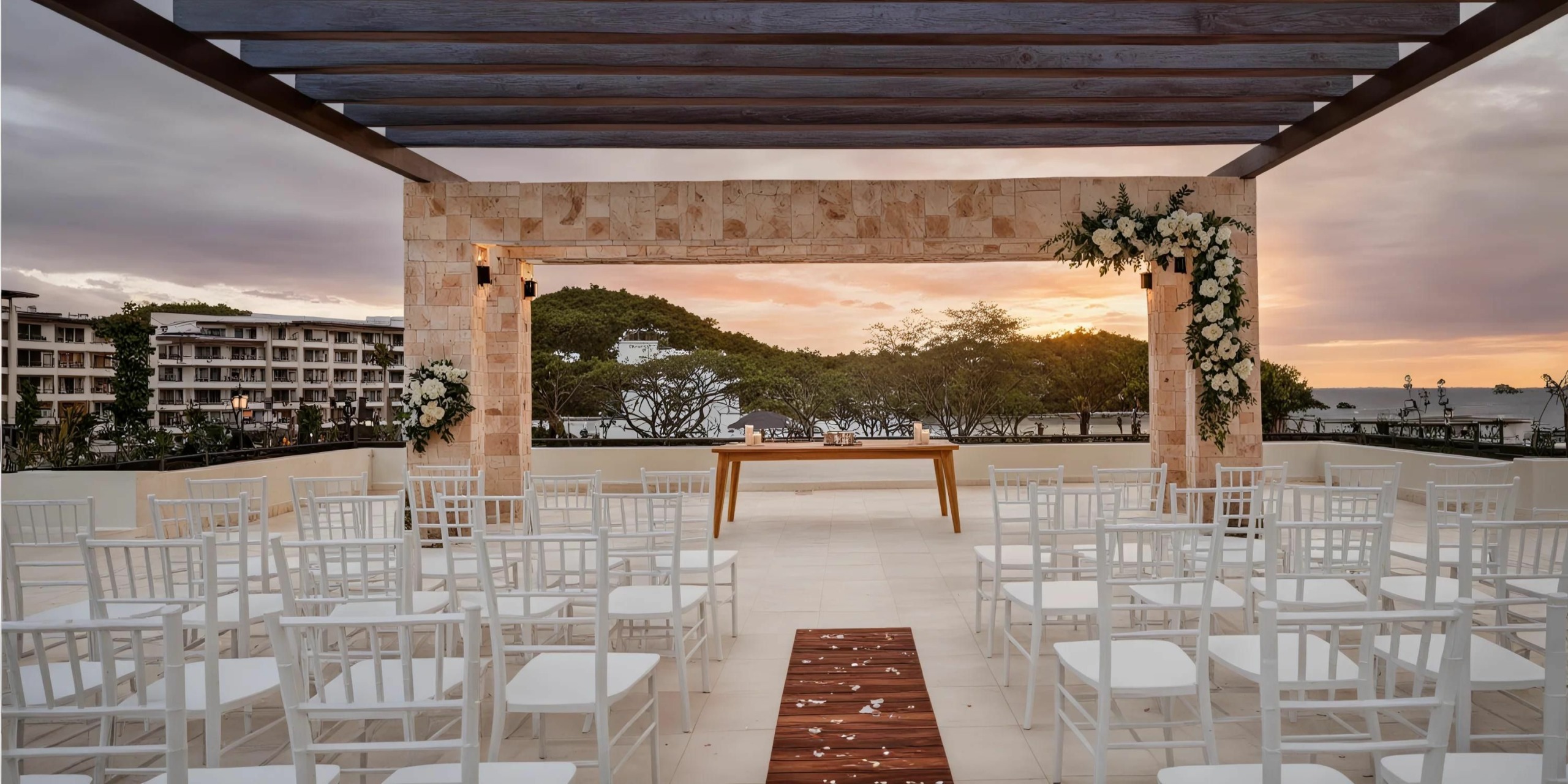Outdoor wedding venue with white chairs arranged in rows, a wooden pathway, and a decorated altar. The background features a sunset, buildings, and trees.