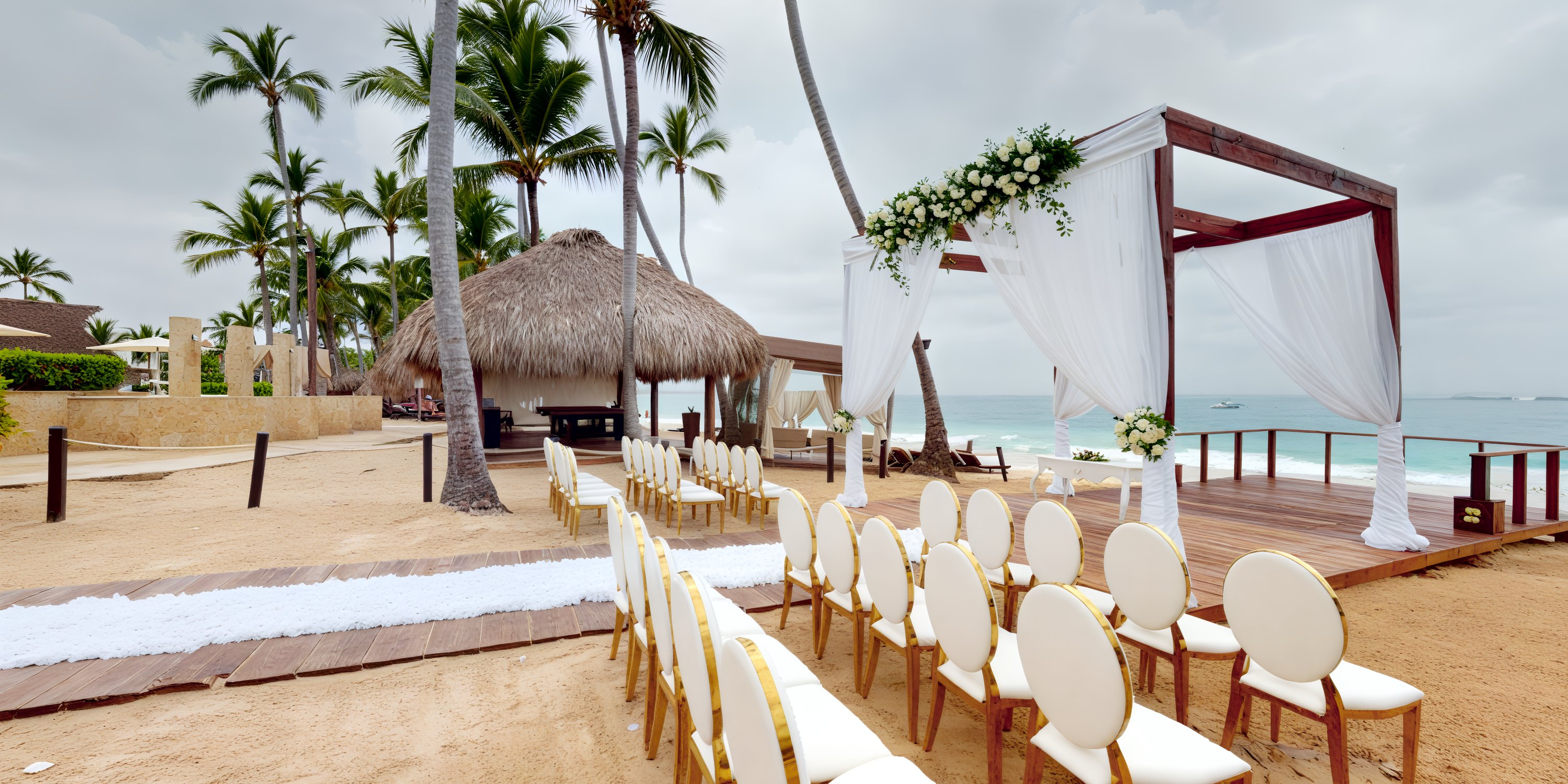 A destination wedding setup on the beach features rows of chairs oriented towards a beautifully decorated archway, with a charming thatched-roof hut situated nearby.