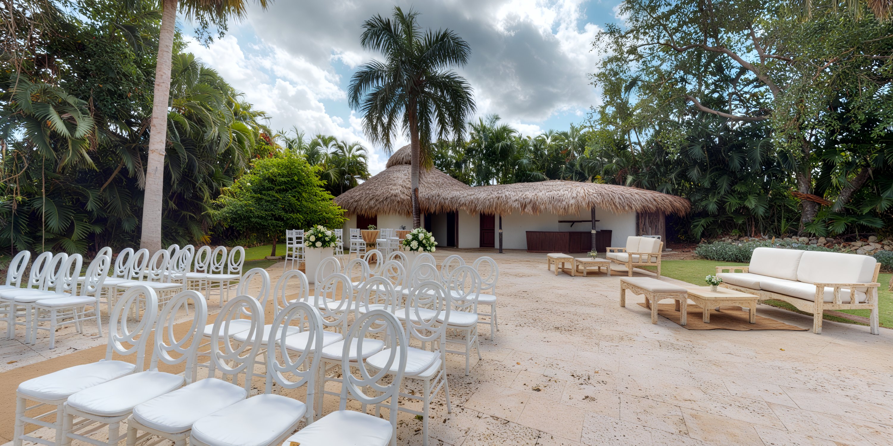 A destination wedding setup featuring white chairs, surrounded by tropical trees and complemented by a thatched roof structure.