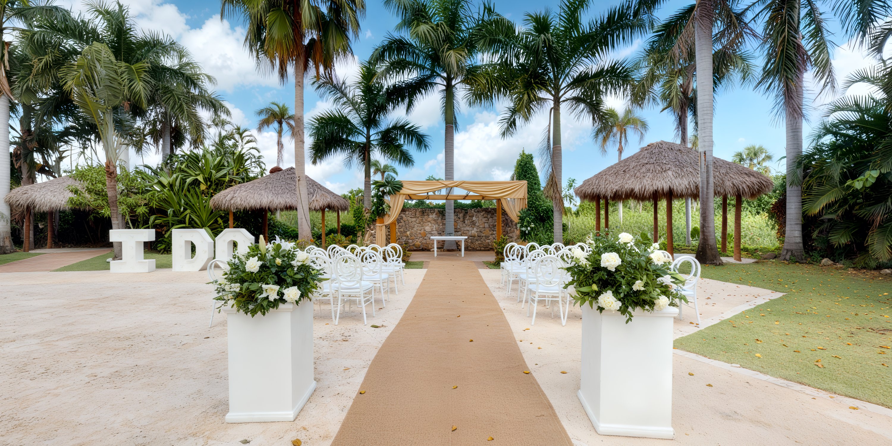 A beautiful destination wedding setup featuring white chairs and exquisite floral arrangements creates a stunning scene under the palm trees, with an enchanting "I DO" sign completing the picturesque backdrop.