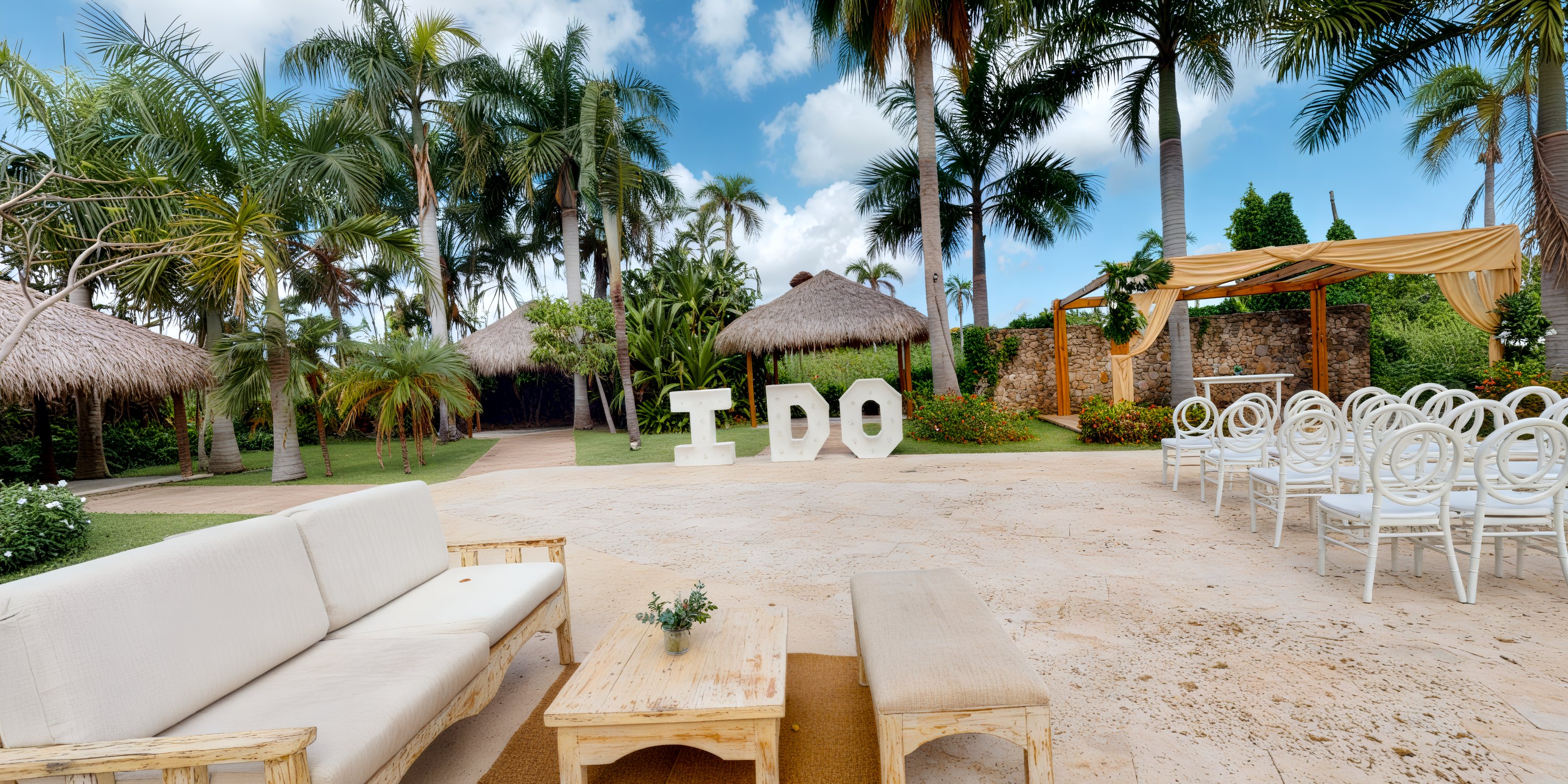 A destination wedding arranged outdoors features a charming "I DO" sign surrounded by tropical palm trees and elegant white chairs.