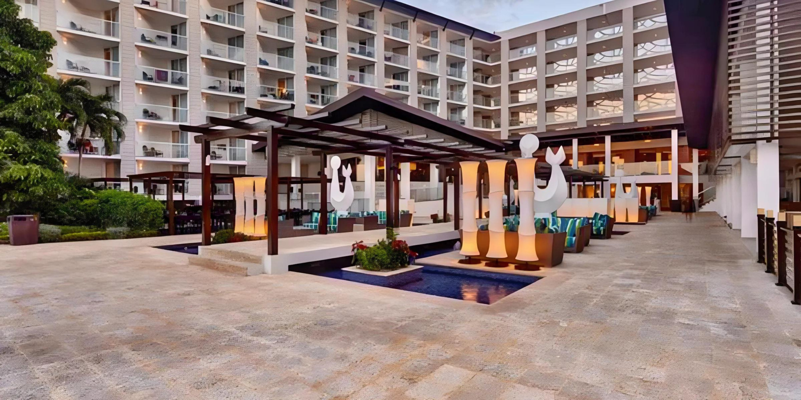 Modern hotel courtyard, ideal for destination weddings, with a covered walkway, seating areas, and large numeral sculptures, surrounded by a multi-story building at dusk.