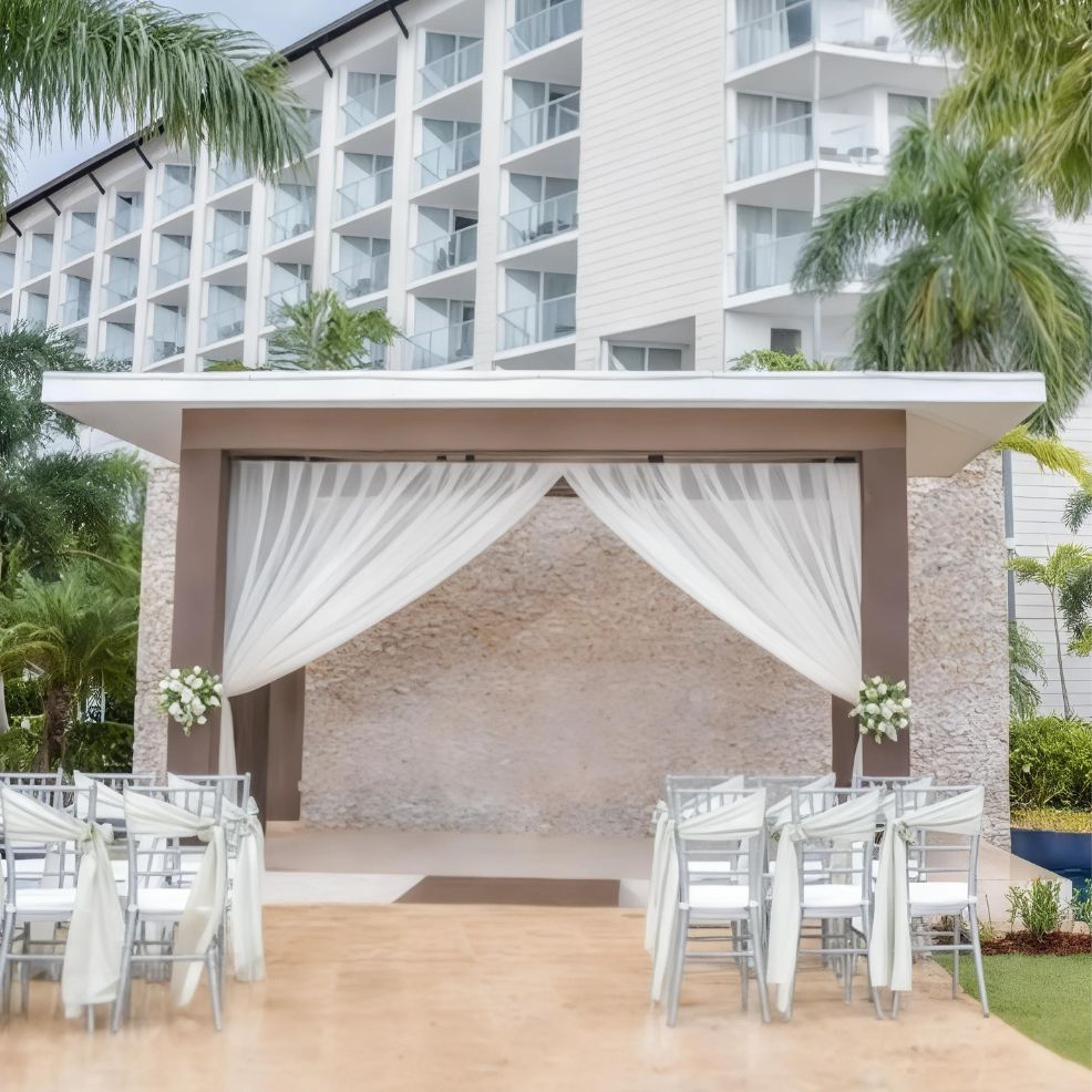 An outdoor wedding venue set up with white chairs, a covered pergola draped with white fabric, and a backdrop of a modern multi-story building.