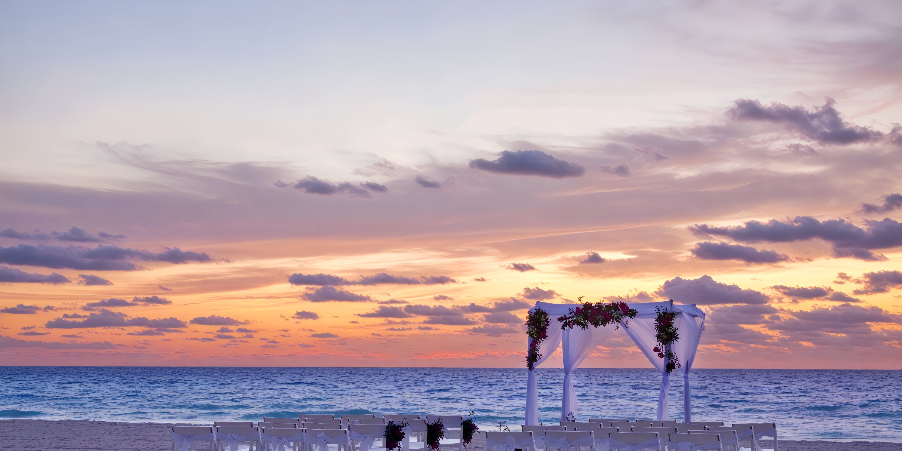 A stunning destination wedding setup at Sandos Cancun features an elegant arch and rows of chairs on the beach, all beautifully illuminated by the setting sun.