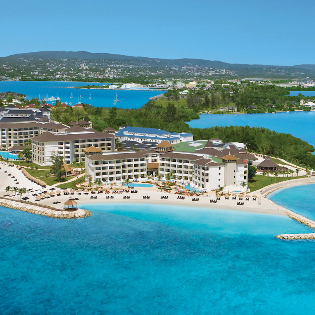 Aerial view of Secrets St. James Montego Bay, popular for destination weddings, featuring white buildings, swimming pools, and a crowded beach overlooking a clear blue ocean and lush green hills in the background.