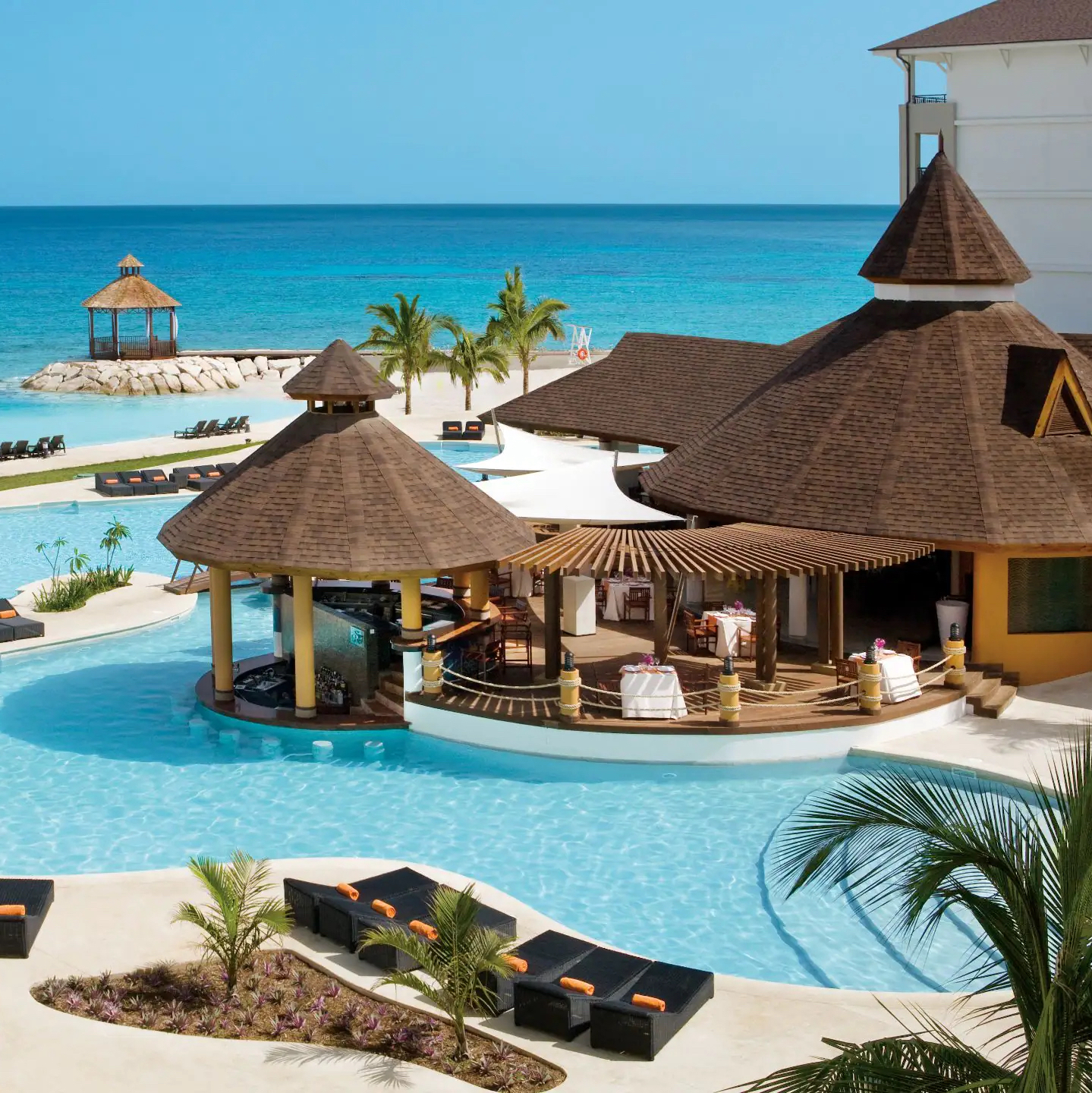 A luxury beachside resort with a circular pool bar under thatched roofs, surrounded by palm trees and lounge chairs, overlooking a serene ocean, perfect for a destination wedding at Secrets Wild Orchid.