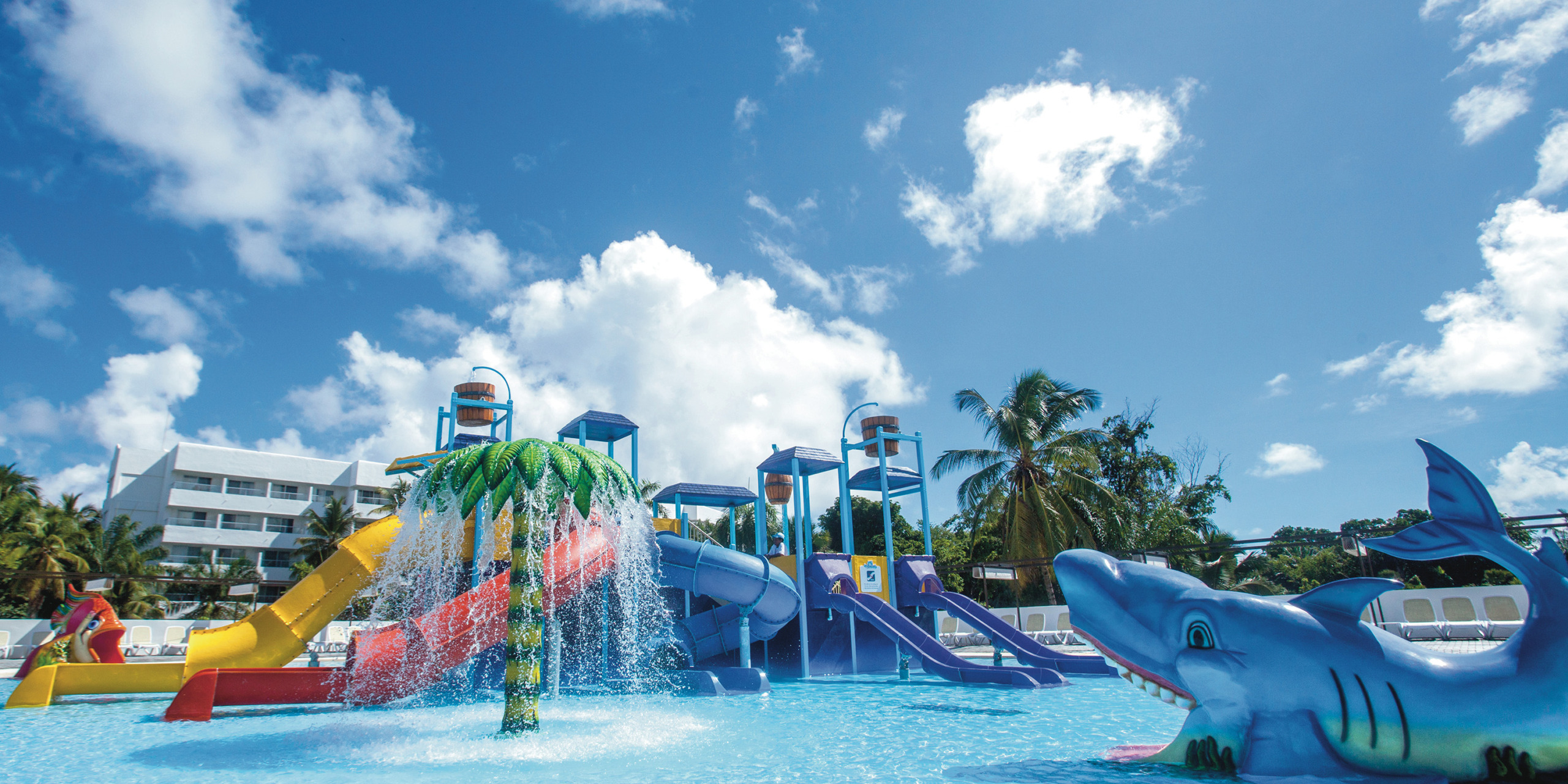 Experience the vibrant Hotel Riu Palace Bavaro, featuring a water park with exhilarating slides, an exciting splash pad, and a striking shark sculpture all set against the backdrop of a clear blue sky.
