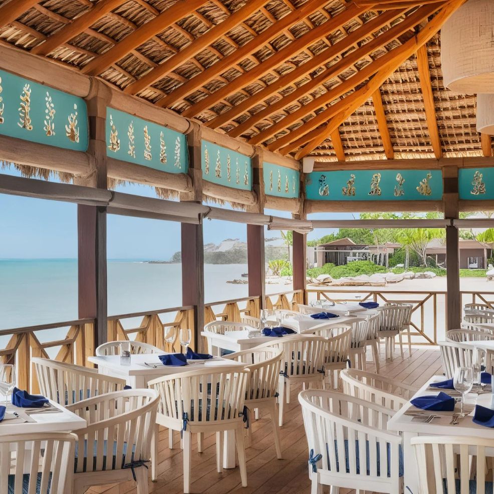 Outdoor dining area with white wooden tables and chairs under a thatched roof, overlooking a sandy beach and calm sea. Blue patterned decor adorns the upper walls. Napkins are neatly placed on tables.