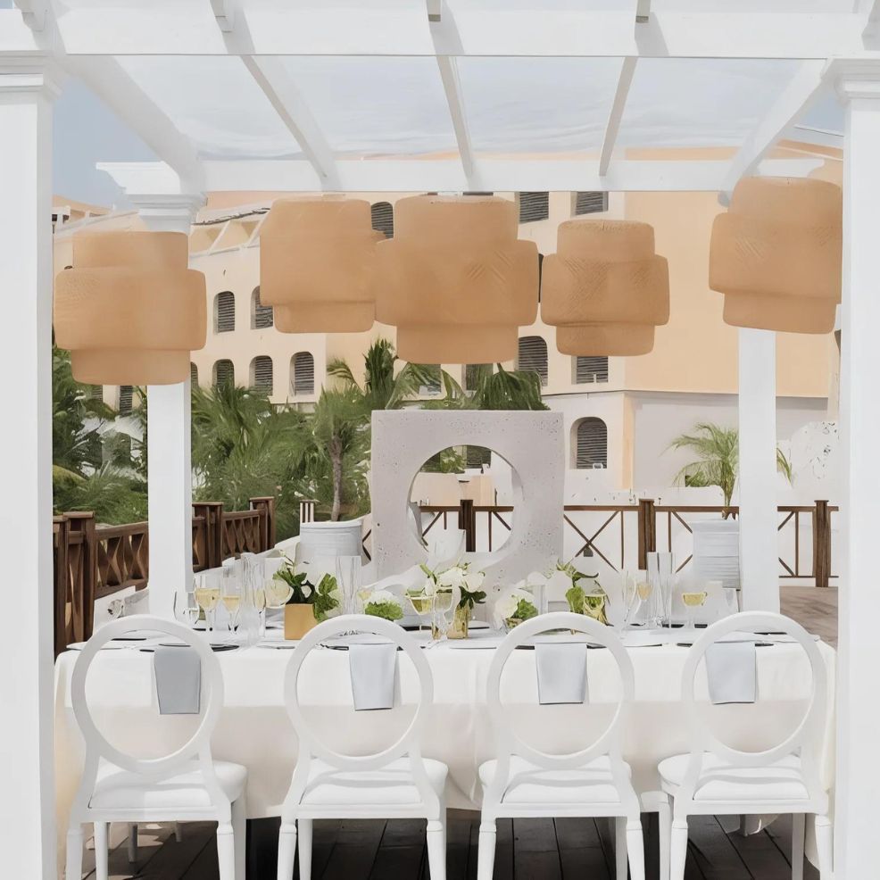 A long, white dining table with six chairs is set for a meal under a pergola with beige lanterns. In the background are palm trees, an ornamental stone structure, and a peach-colored building.