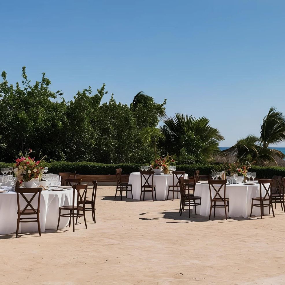 Outdoor event setup with round tables covered with white tablecloths, wooden chairs, and floral centerpieces under a clear blue sky, surrounded by greenery.