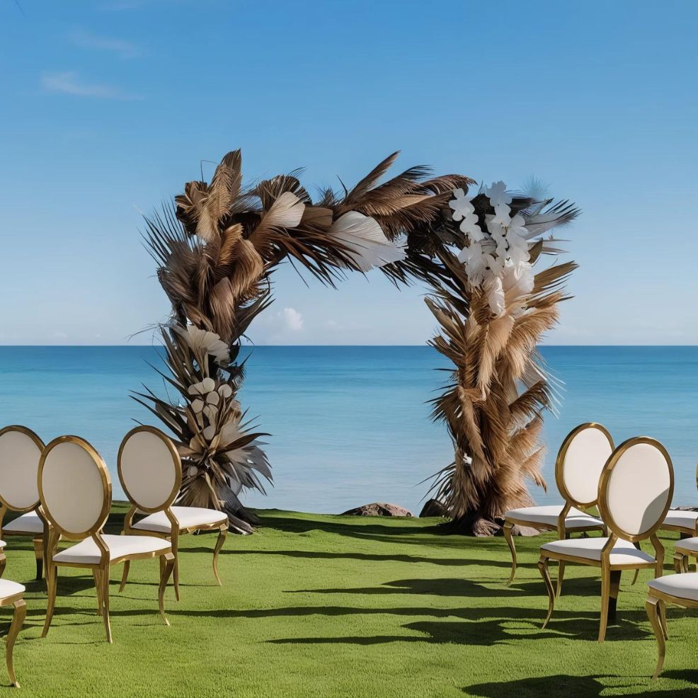 Outdoor wedding arch made of dried palm leaves and white flowers, set on green grass with gold-trimmed chairs. Background shows a calm ocean under a clear blue sky.