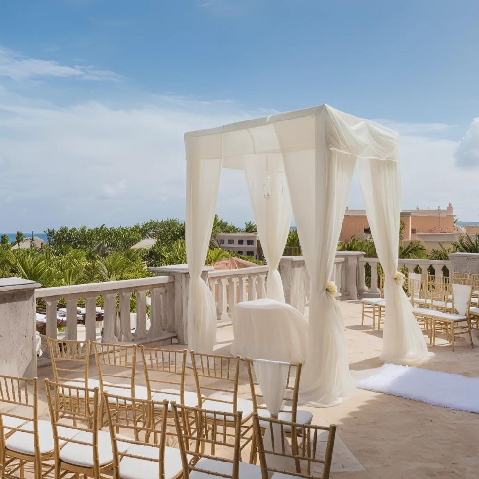 An outdoor wedding ceremony setup with a white canopy and rows of gold chairs on a terrace overlooking a scenic landscape with greenery and buildings under a blue sky.