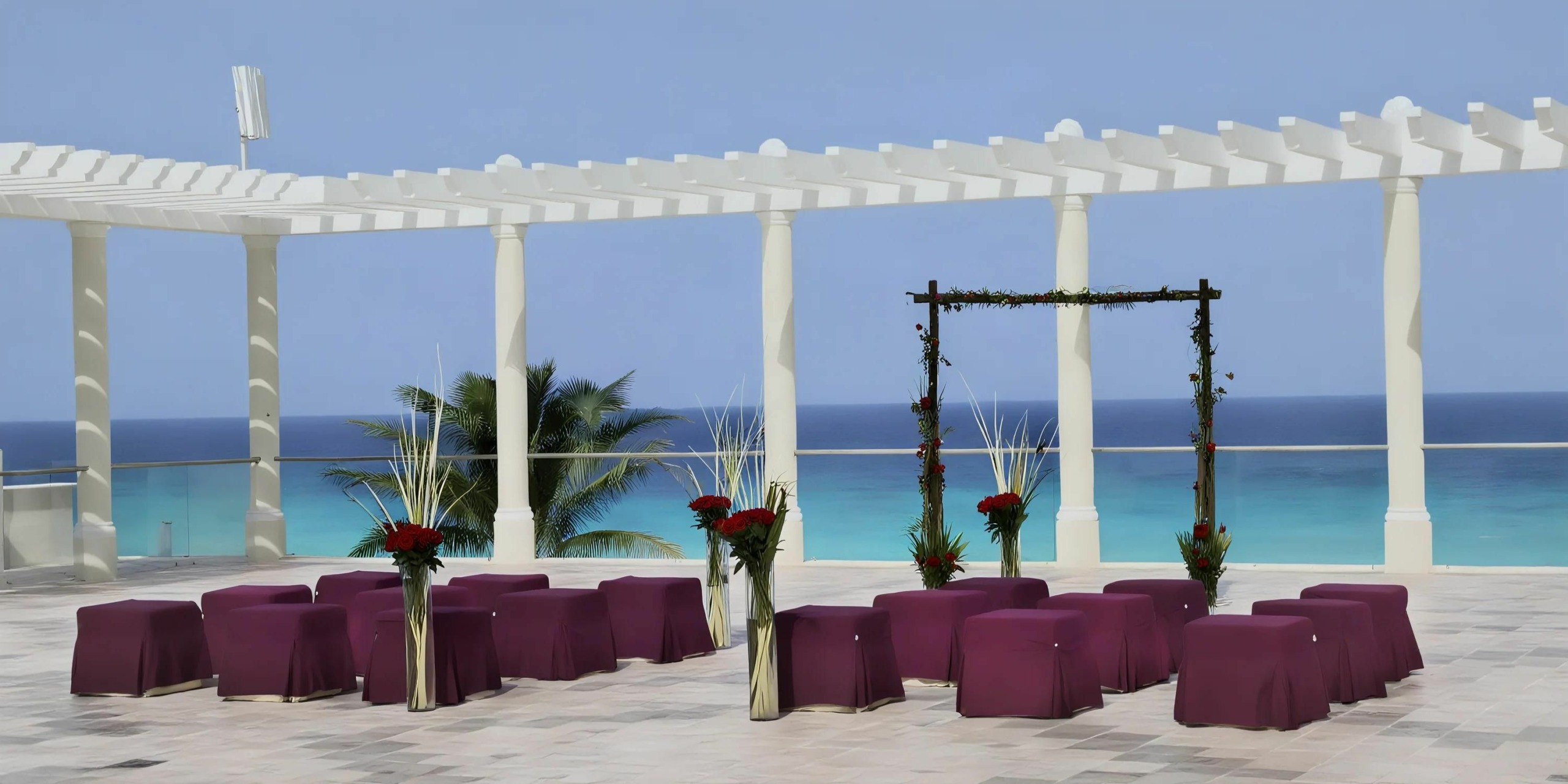 An outdoor wedding setup with purple-covered chairs arranged in rows facing a decorated arch under a white pergola, overlooking the ocean.