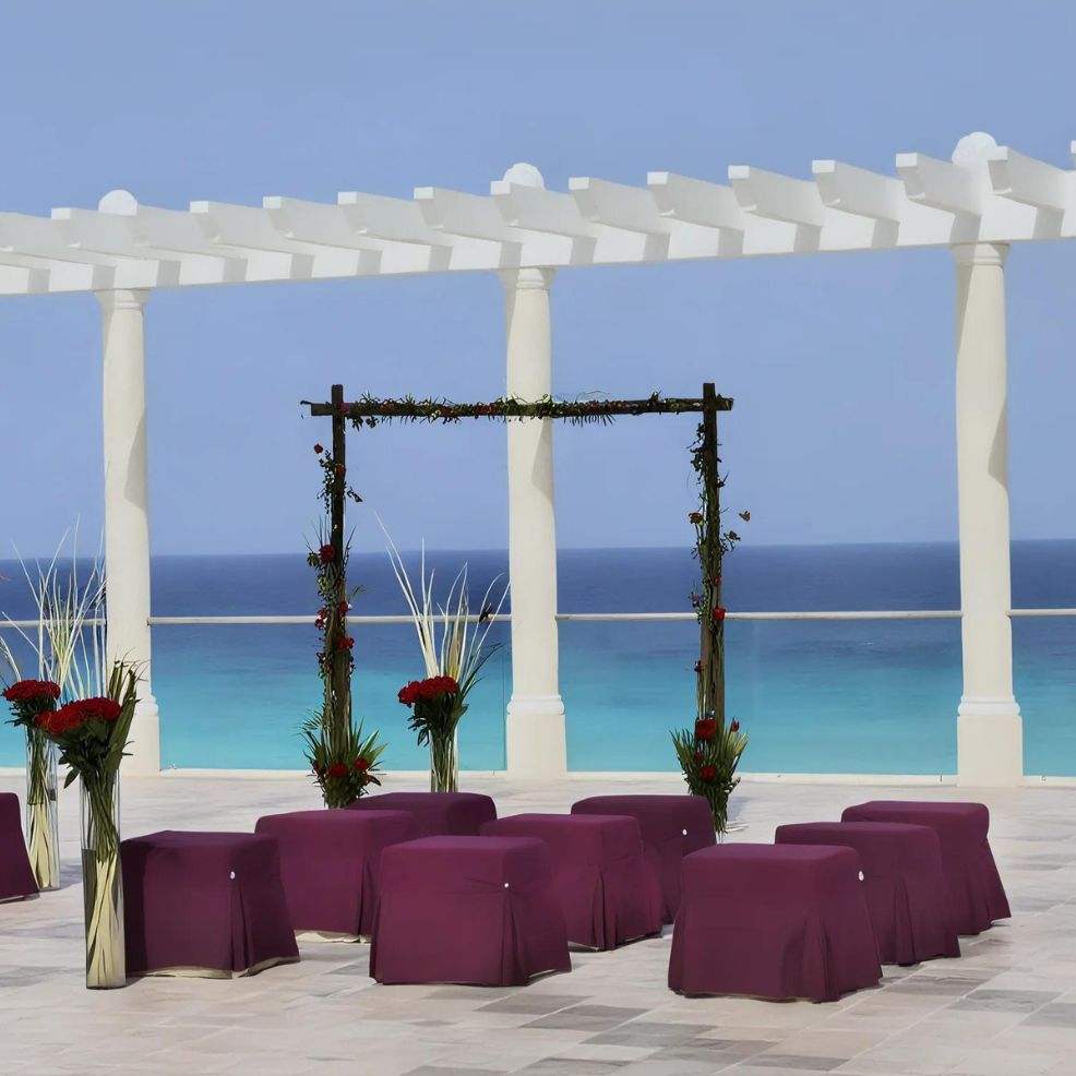 A wedding altar setup with red-covered chairs, floral arrangements, and a canopy overlooking a clear blue ocean under a pergola.