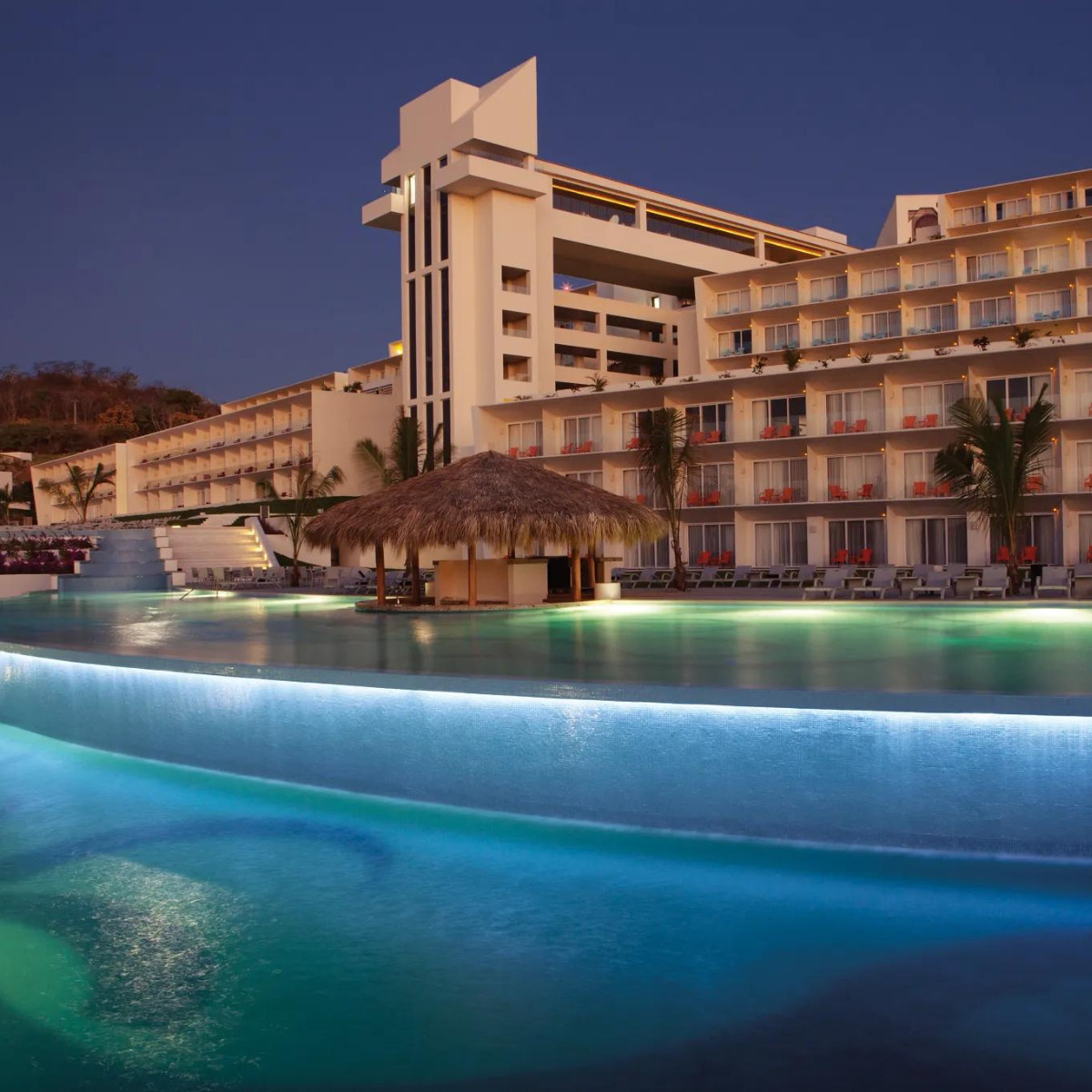 An illuminated pool in the evening, curving in front of Secrets Huatulco with illuminated balconies and palm-thatched umbrellas, perfect for a destination wedding.