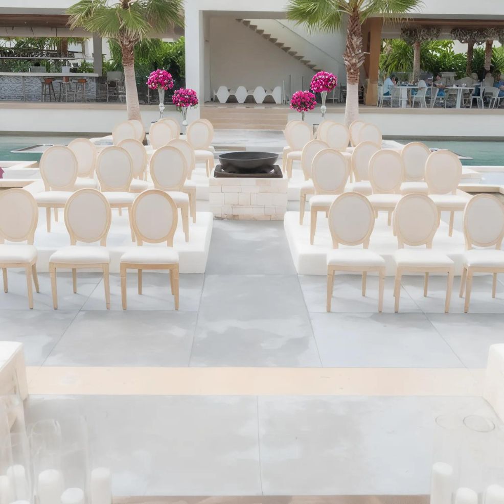 The outdoor venue at Secrets Riviera Cancun features two rows of white chairs facing a modern altar adorned with a black bowl centerpiece, flanked by potted pink flowers. Palm trees provide a picturesque backdrop, and a covered area with additional seating completes the setting.