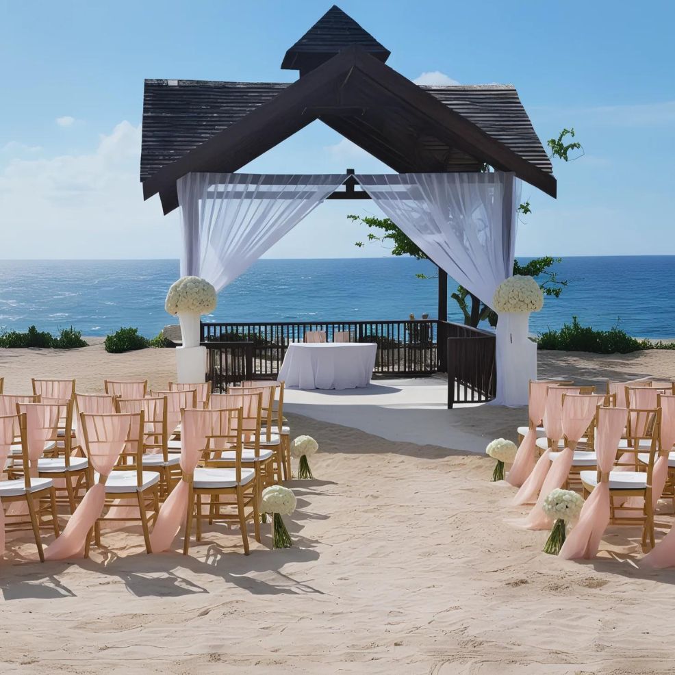 An outdoor beach wedding setup with a wooden arch and white drapes, an aisle lined with wooden chairs draped in pink fabric, and a table under the arch, overlooking the ocean.